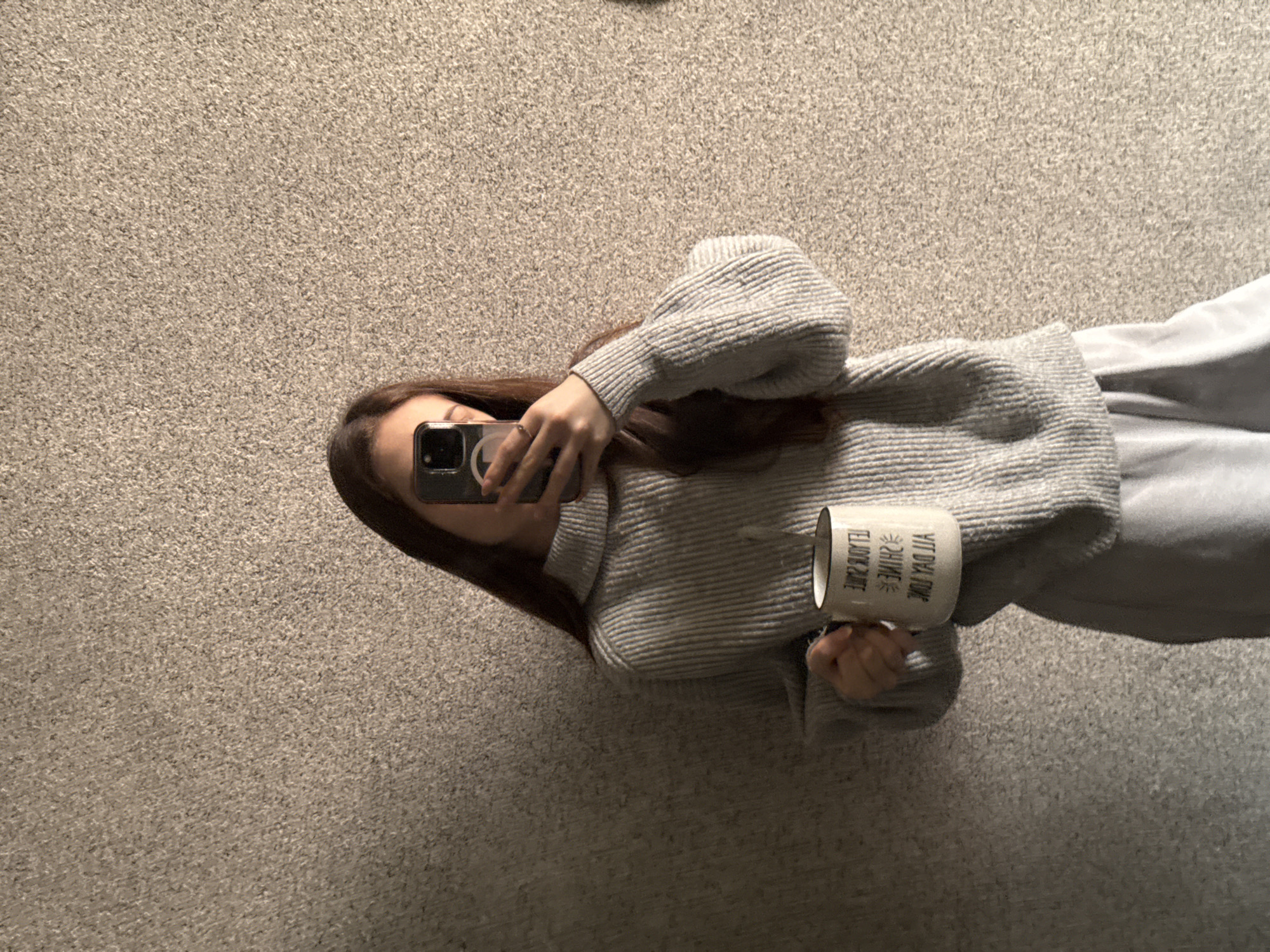 A person is standing on a carpet, holding a mug in one hand and a phone in the other, taking a picture of themself. The person is wearing a grey sweater and grey pants. The mug has writing on it. The background is a beige carpet. The location is unknown, but it could not be determined that the location is Sofia, Bulgaria.