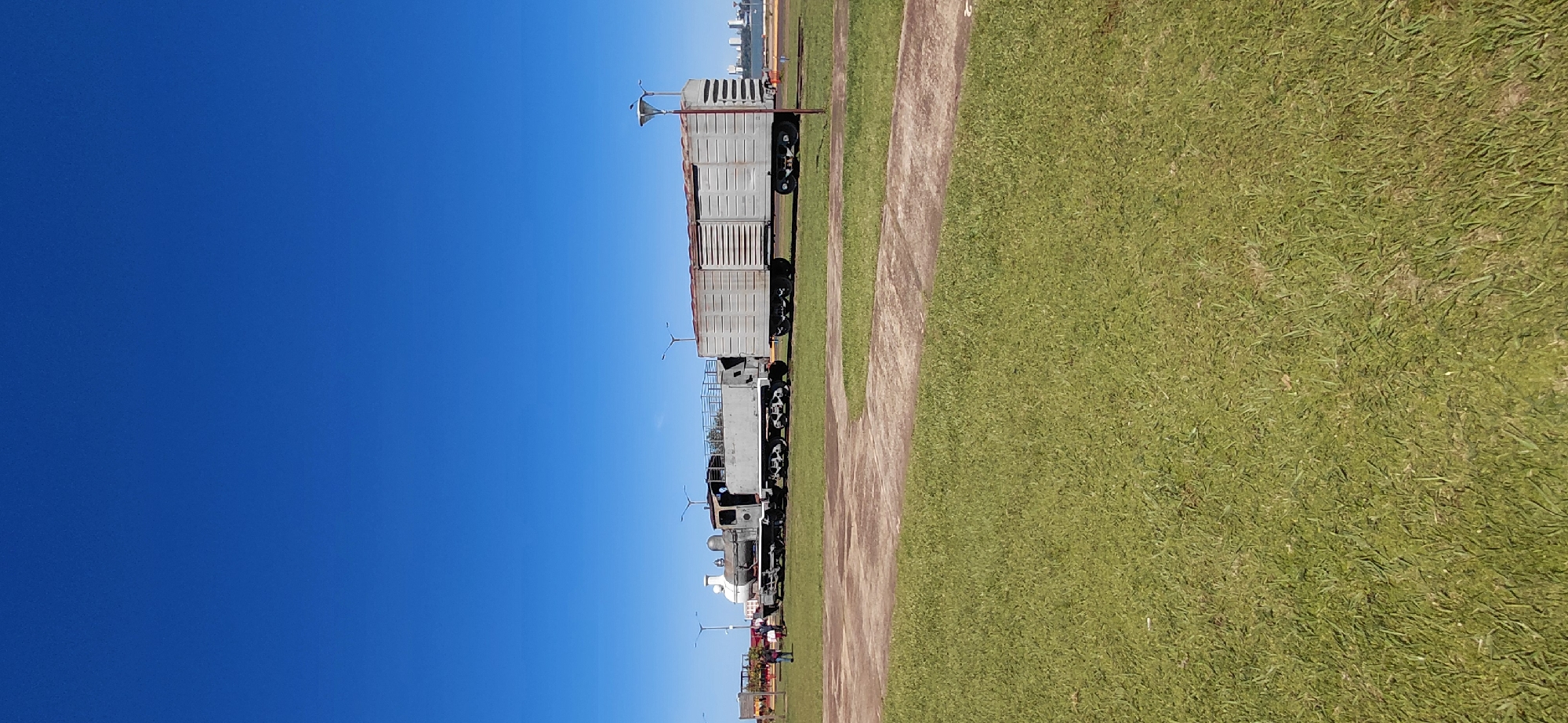 The image depicts a historic train on display in a park in Encarnacion, Paraguay, with a clear blue sky overhead. The train consists of a locomotive and at least one freight car. The locomotive appears to be a steam engine, painted in gray and white. The freight car is also gray and has a corrugated metal exterior with visible siding. The train is situated on tracks in a grassy area, with a concrete path curving through the foreground. In the background, several lampposts are visible, and beyond that, a body of water can be seen, with buildings and a cityscape on the far shore, indicating the presence of a river or bay. A few people are standing near the locomotive, and what appears to be an exhibit or playground structure is visible to the far left. The overall scene suggests a well-maintained public space.