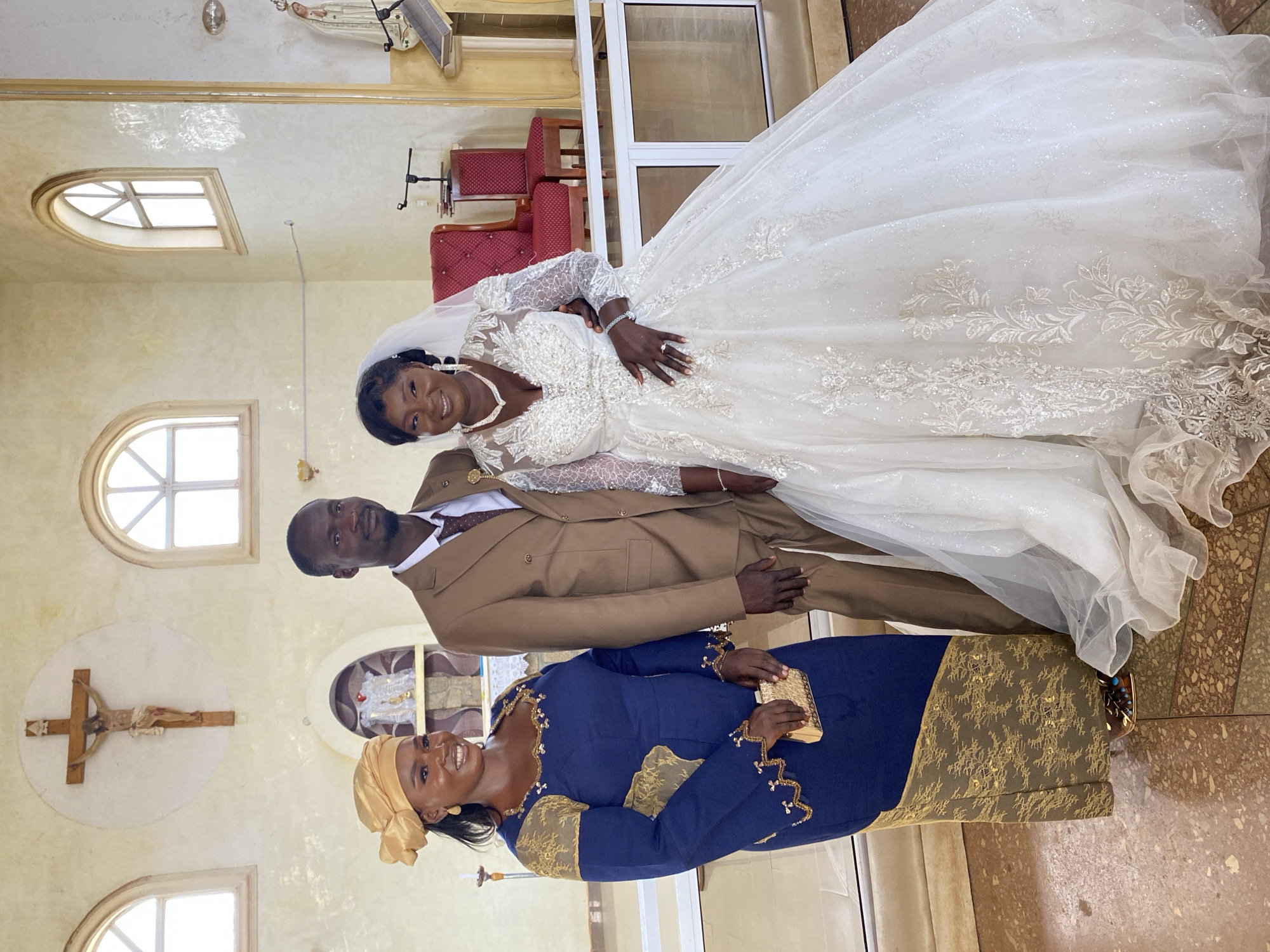 Three people stand for a photograph in what appears to be a church in Kuru Karama, Nigeria.
On the right, a woman in a sparkling white wedding gown with long lace sleeves, a veil, and jewelry poses with her hand on the man beside her.
The man, dressed in a brown suit, stands next to her.
In the foreground, a woman wears a blue dress with gold detailing and a matching head covering. She holds a gold clutch.
Above, a crucifix hangs on the wall. Arched windows provide light. In the background, church chairs are visible behind a doorway. The floor is tiled.