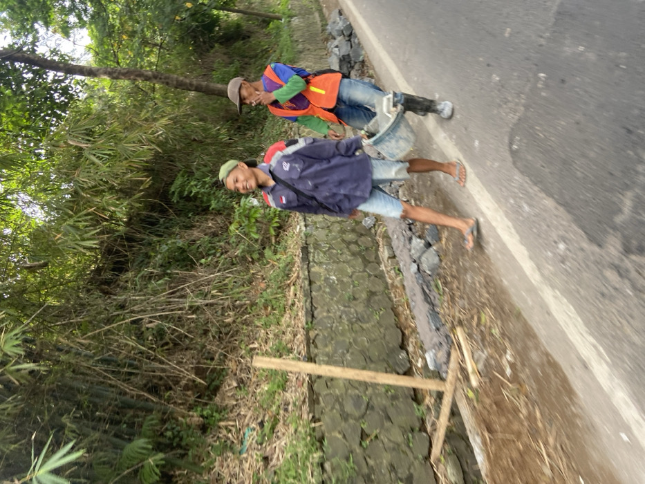 Two men are walking along a paved road in Soreang, Indonesia. The man on the left is wearing a blue shirt, blue jeans, a green cap, and sandals. He is smiling and looking to the left. The man on the right is wearing a green and orange safety vest, blue jeans, and a brown cap. He is carrying a bucket, and looking down. There are several rocks on the shoulder of the road and a stone wall behind the men. The men are walking towards the right side of the image. There are trees and foliage on the left side of the image. The road is lined with a white line. The location of the image is a paved road with a stone wall and trees on the left side, and rocks on the shoulder of the road. The coordinates provided indicate a location in Soreang, Indonesia.