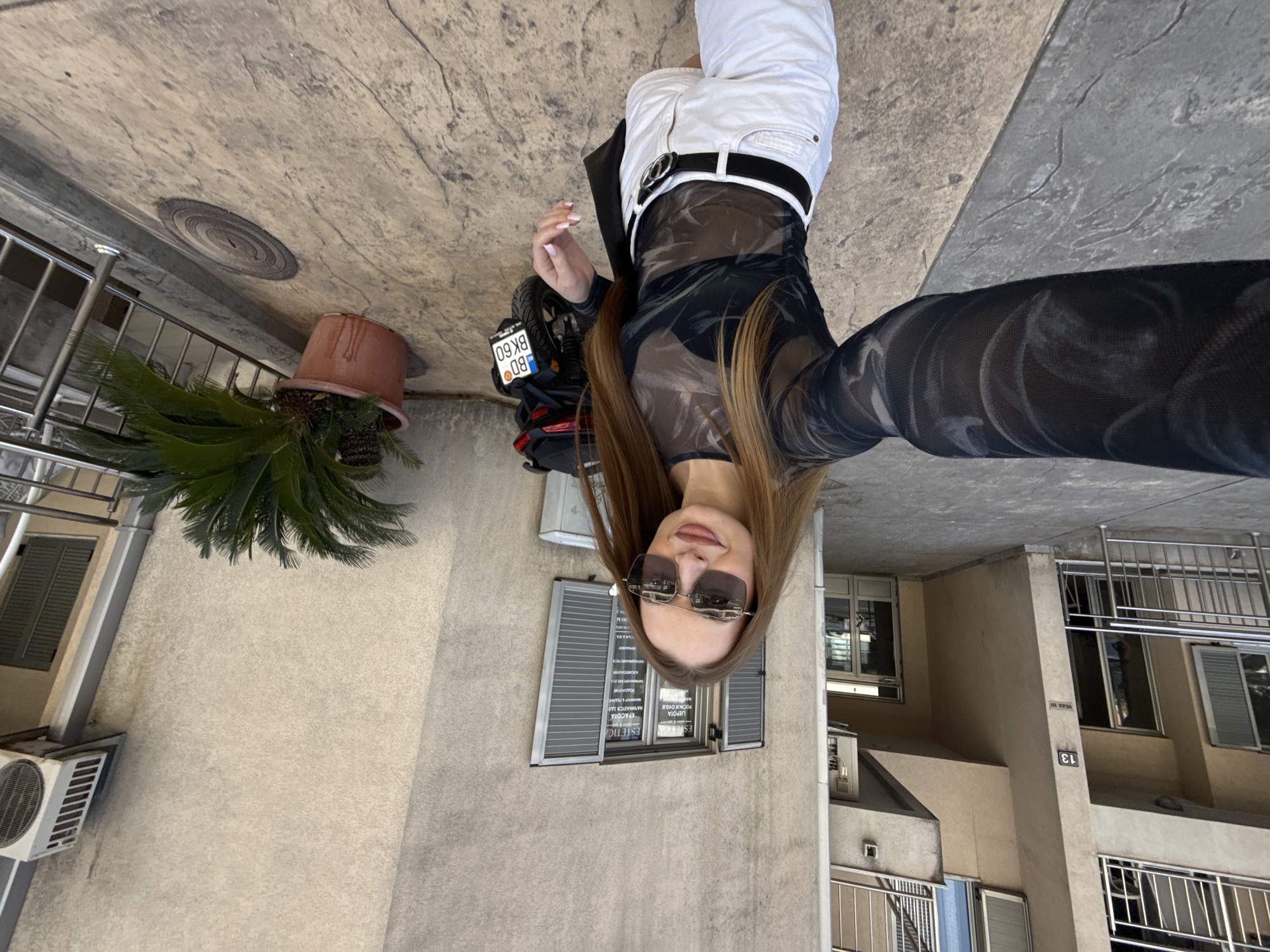 This media file captures a young woman taking a selfie outdoors in a daytime setting, likely in Budva, Montenegro. The image is oriented vertically, with the woman in the foreground and urban surroundings behind her.
The central subject is a young woman with long, light brown hair, wearing large dark sunglasses. She is dressed in white shorts, a black belt with a silver buckle, and a sheer black long-sleeved top featuring a subtle, swirling pattern. Her right hand, partially visible, has light pink nail polish.
To her left, a black motorcycle or scooter is parked, with its license plate clearly showing "BD 60" followed by obscured characters, confirming the Budva location (BD is the vehicle registration code for Budva). Adjacent to the motorcycle is a large potted plant, resembling a palm, contained within a terracotta pot.
In the background, multi-story buildings with light-colored, possibly beige, facades are visible. These buildings feature multiple windows, some with closed gray shutters, and balconies equipped with metal railings. An air conditioning unit is mounted on one of the lower walls. On a window directly behind the woman, reversed text from inside a business is readable, when correctly oriented, as "ESTETIC" followed by various Serbian/Montenegrin words related to beauty and wellness services, such as "LEPOTA I ZDRAVLJE" (Beauty and Health), "MASAŽE" (Massages), "DEPILACIJA" (Depilation), and other treatments. A building number "13" is also visible on a structural pillar.
The scene is brightly lit, indicating clear, sunny weather. The ground beneath the woman is a textured concrete or paved surface.