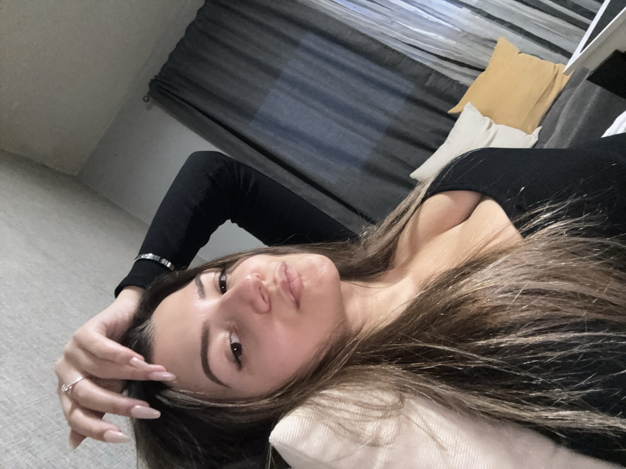 A woman is lying down indoors, facing the camera. Her head rests on a light-colored pillow. She wears a black long-sleeved top. She has brown hair and light skin. Her left hand is raised near her face, with a ring visible on one finger. Behind her is a sofa with a yellow pillow and a white pillow. Grey curtains are in the background. The location is unknown, but assumed to be in Sofia, Bulgaria.