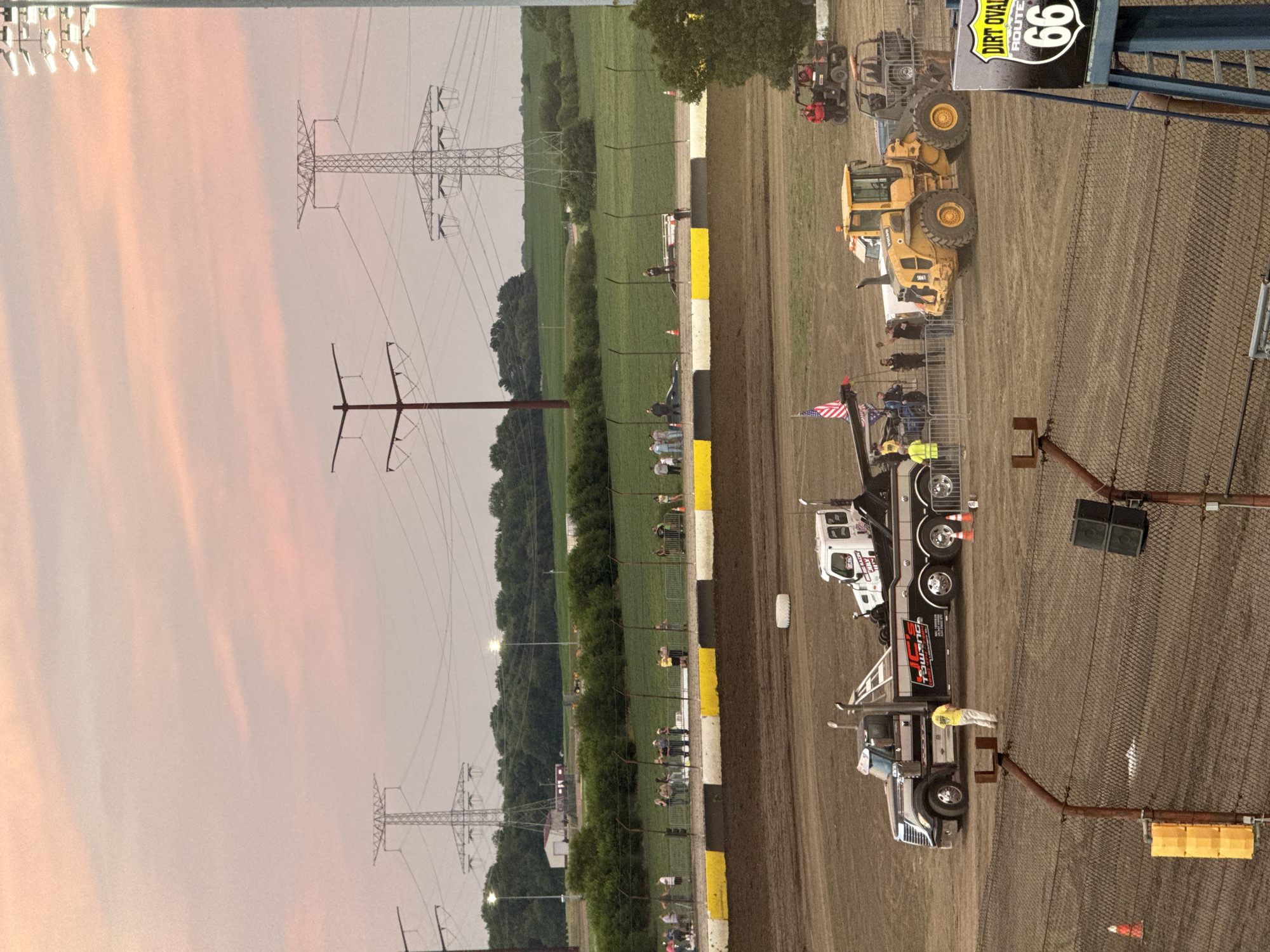 The image captures a dirt oval race track in Joliet, Illinois, during dusk. The sky is painted with soft pink and orange hues from the setting sun, with prominent power lines and transmission towers stretching across the horizon. In the foreground, a tow truck is parked on the dirt track, and nearby, a yellow front-end loader and a red ATV are also present. Several people are seen on and around the vehicles, some appearing to be working or observing the scene. A fence separates the track from a spectator area where a group of people are gathered. In the distance, lush green fields and trees form the backdrop, suggesting a rural or semi-rural setting on the outskirts of Joliet. A sign for "Dirt Oval Route 66" is visible on the right side of the image, identifying the location.