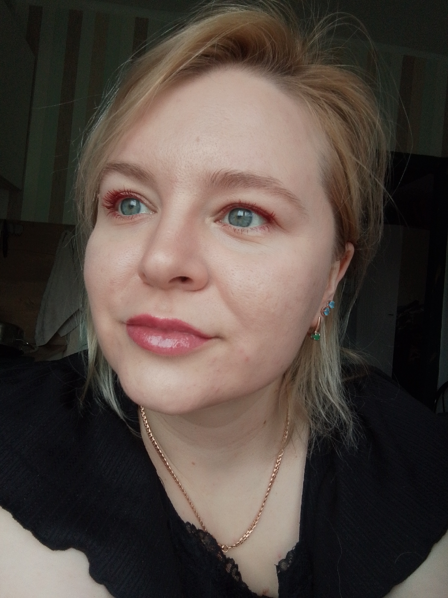 A medium close-up image depicts a fair-skinned person with blonde hair, fair eyelashes, and a light pink lip gloss. The person is wearing what appears to be a black top with lace detail and a gold chain necklace. One earring is visible; it is a gold, vertical earring with blue and green stones. The background is blurred, showing what appears to be a striped wall and a glimpse of a room. No elements indicate the exact location in Izhevsk, Russia.