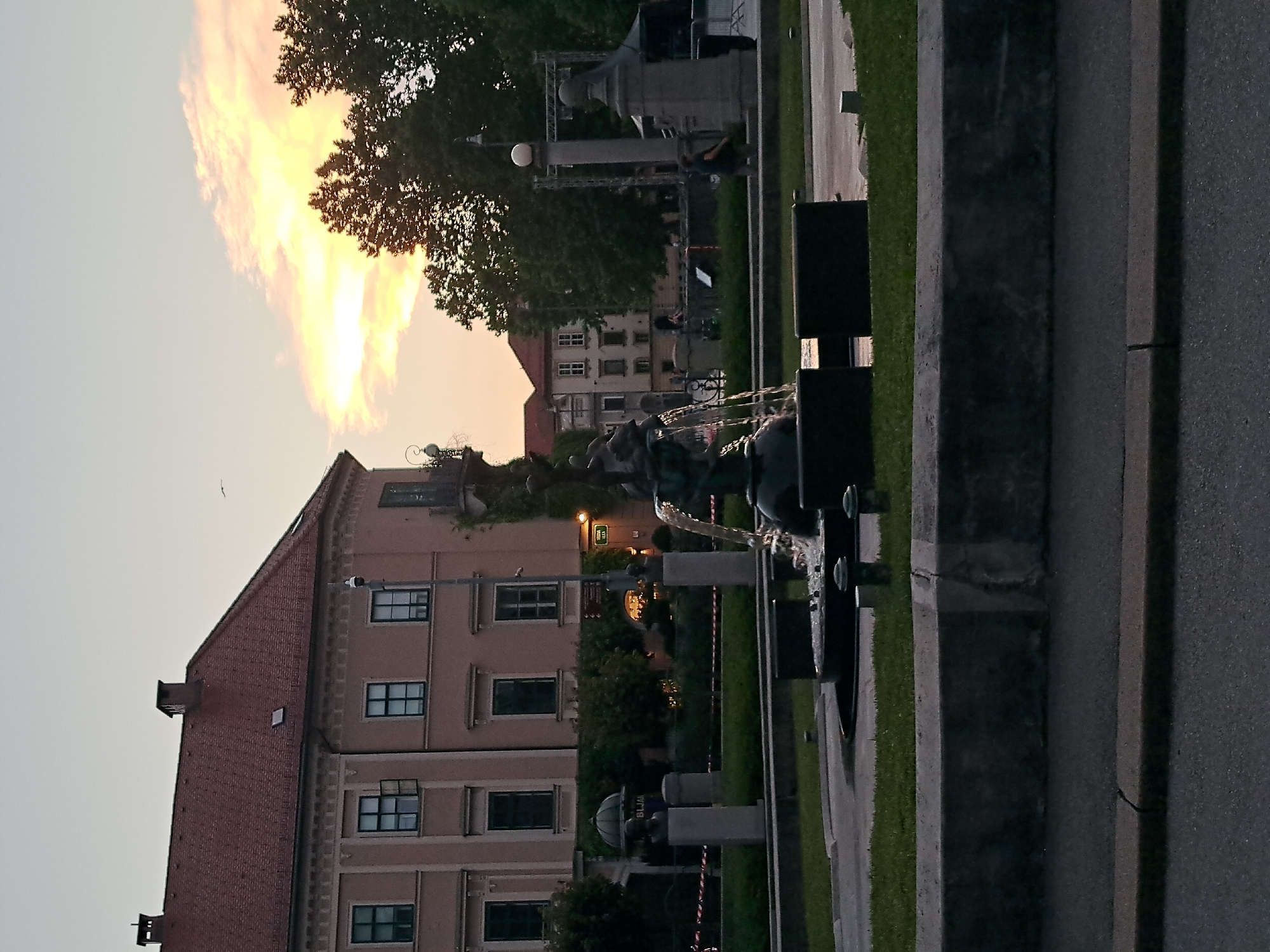 The image shows a public square with a fountain in the foreground. The fountain has a sculpture of figures on top and water is flowing from it. In the background, there is a large building with a red tiled roof and several windows. There are also trees and streetlights. The sky is visible in the upper right corner and appears to be sunset with some clouds. There are some people in the background, but they are not clearly visible. The overall scene suggests a peaceful urban setting. The location is in the city of Unknown, Unknown.