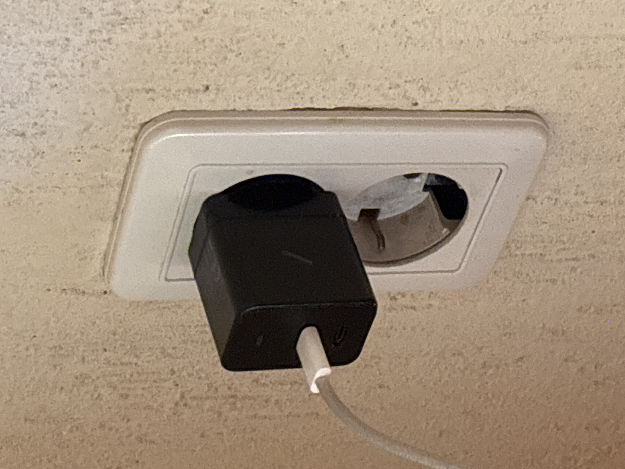 The image shows a close-up of a white electrical outlet on a textured, beige wall. A black power adapter, likely for a mobile device, is plugged into the top receptacle of the outlet. A white charging cable extends from the adapter. The bottom receptacle of the outlet is empty. The overall scene suggests a typical indoor setting, possibly a residential or hotel room in Krasnaya Polyana, Russia, given the outlet type which is common in the region.