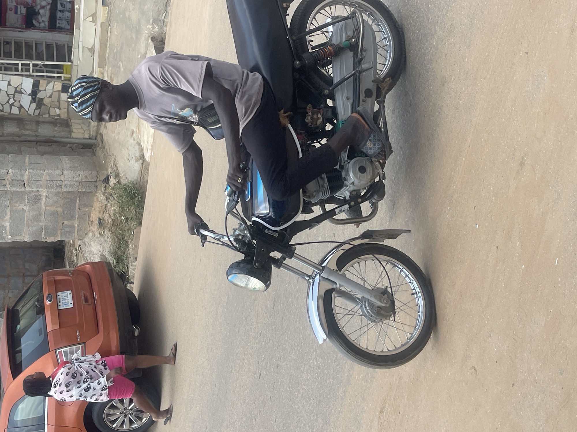 The image shows a young Black man riding a motorcycle in what appears to be a residential or commercial area in Abuja, Nigeria. He is wearing a grey t-shirt, black trousers, and a blue and black striped headwrap. He has a ring on his left hand. The motorcycle is silver and black, with a visible engine and exhaust pipe. The front wheel of the motorcycle is prominent in the foreground, with a round headlight.
In the background, to the left of the motorcycle, is an orange SUV. A young Black woman is standing next to the rear of the SUV, facing away from the camera. She is wearing a white and black patterned top and pink shorts. She is also wearing flip-flops.
The scene is set on a paved surface, likely a road or sidewalk. The buildings in the background are made of brick and stone. The lighting suggests it is daytime, with no clear indication of the specific time or weather conditions. The overall atmosphere seems casual and everyday.
There is visible text on the license plate of the orange SUV, which reads "K1916-AB". There is also some text on the man's t-shirt, which is difficult to read but appears to be a graphic design with some lettering.