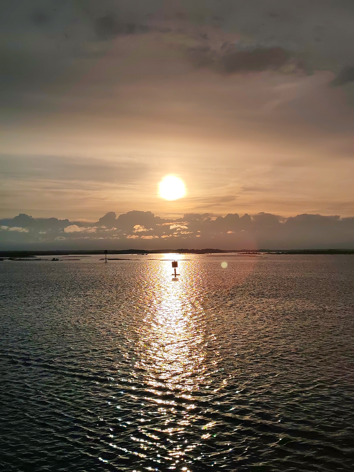 A wide shot captures a serene sunset over water. The sun is a bright orb in the sky, partially obscured by clouds. Its reflection stretches across the water, creating a shimmering path of light. In the distance, land is visible, dotted with what appears to be navigational markers or buoys. The sky transitions from a bright, warm orange near the sun to darker, gray clouds overhead. The water has gentle ripples. The location is not identifiable as Bereg, Russia, from the image alone.