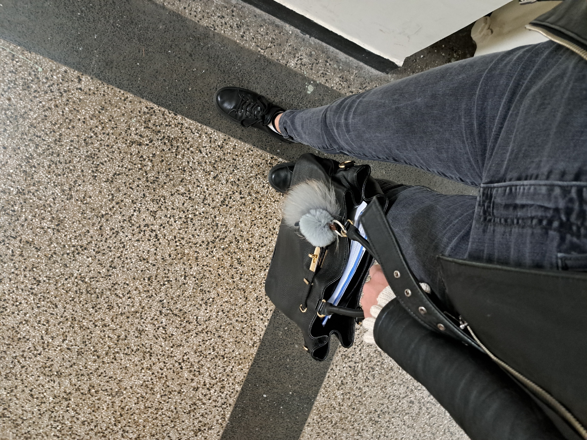 A high-angle shot shows a person standing on a patterned floor with a dark stripe, potentially inside a building. The person is wearing grey jeans, black shoes and a black leather jacket. A black bag with a grey pom-pom and gold hardware is also visible. The setting looks like a public space, such as a station or shopping mall. Sofia, Bulgaria is not visible or identifiable in the image.