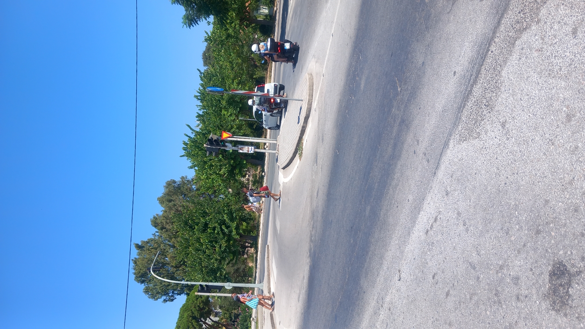 This is a vertical, eye-level, full shot of a street intersection under a clear blue sky in Kos, Greece. The intersection is paved with asphalt and features a pedestrian crossing. In the foreground, the road is visible with the sun casting shadows. On the left side of the intersection, two women in casual summer attire are walking. One woman is wearing a blue and white striped dress, and the other is wearing a pink top and a skirt. They appear to be crossing the street from left to right.
Further into the intersection, a man is walking across the pedestrian crossing from right to left, wearing a dark t-shirt, shorts, and flip-flops, carrying a red bag. To the right of him, a white car is stopped at the intersection. Behind the white car, a black scooter is in motion, with two individuals wearing helmets. The driver of the scooter is a woman, and a passenger is seated behind her.
On the right side of the intersection, another scooter with two individuals wearing helmets is also in motion. The traffic lights are visible on poles, indicating a controlled intersection. Lush green trees line the background of the intersection, with some buildings partially visible behind them. The overall scene depicts a sunny day at a busy intersection in a Mediterranean town.