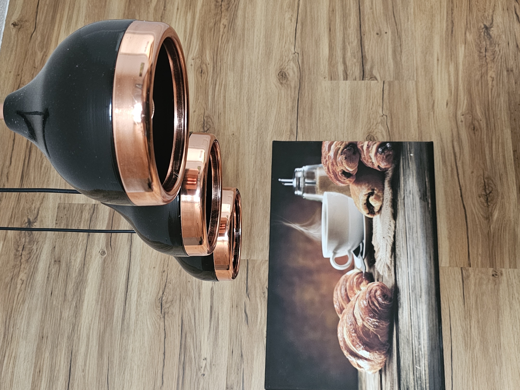 Three black pendant lights with copper trim hang in front of a wood-paneled wall. Below the lights, a framed picture depicts a breakfast scene with croissants, a cup of coffee, and a sugar dispenser on a wooden table. The scene is captured in a close-up, eye-level shot.