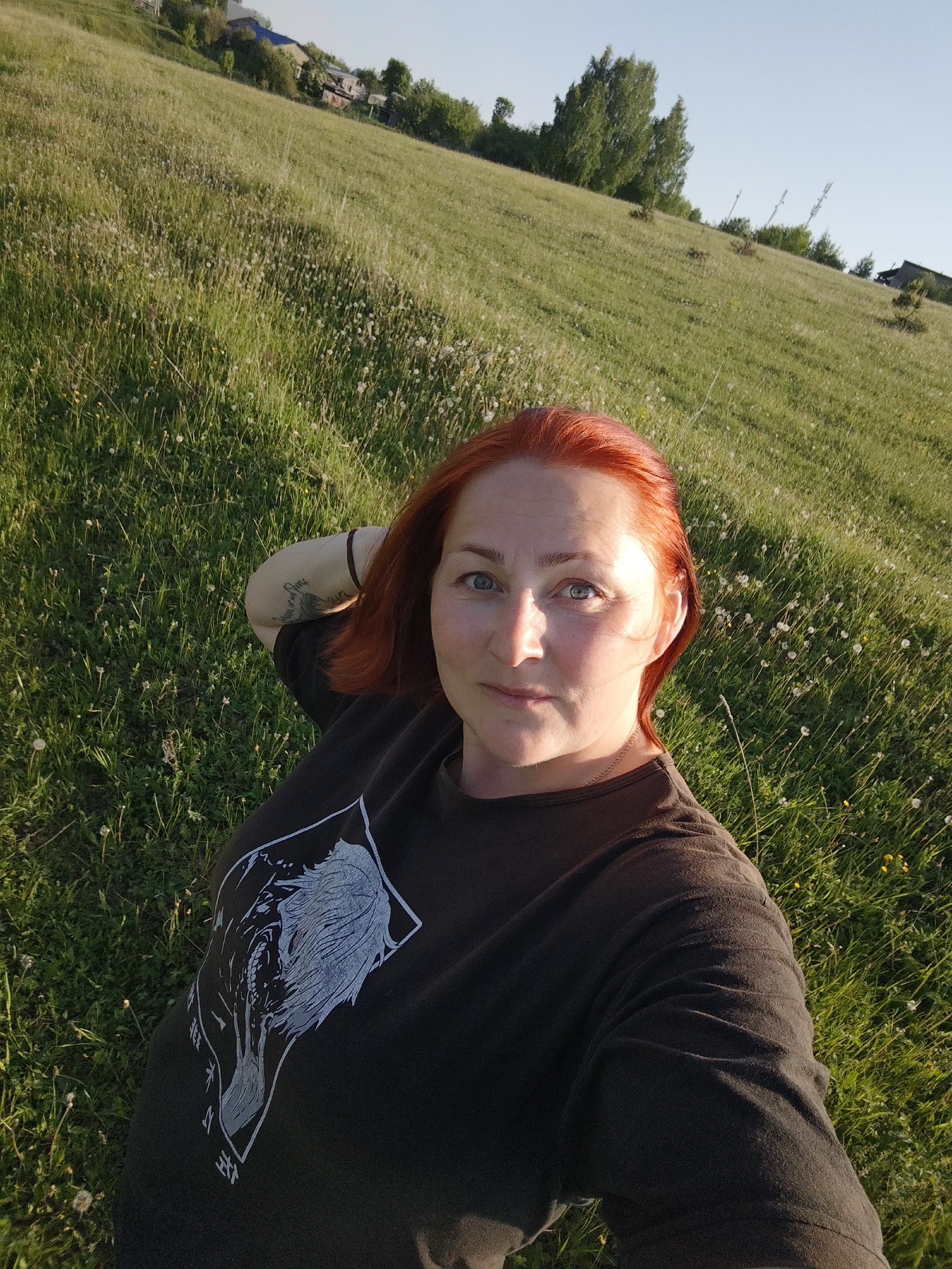 A selfie captures a woman with red hair lying in a grassy field. The field slopes upward into a hill dotted with dandelions and low foliage. In the background, hints of buildings and trees suggest a settled area. The location is Фоки, Russia. The woman wears a dark T-shirt with a graphic print. A tattoo is visible on her arm. The lighting indicates it is daytime.