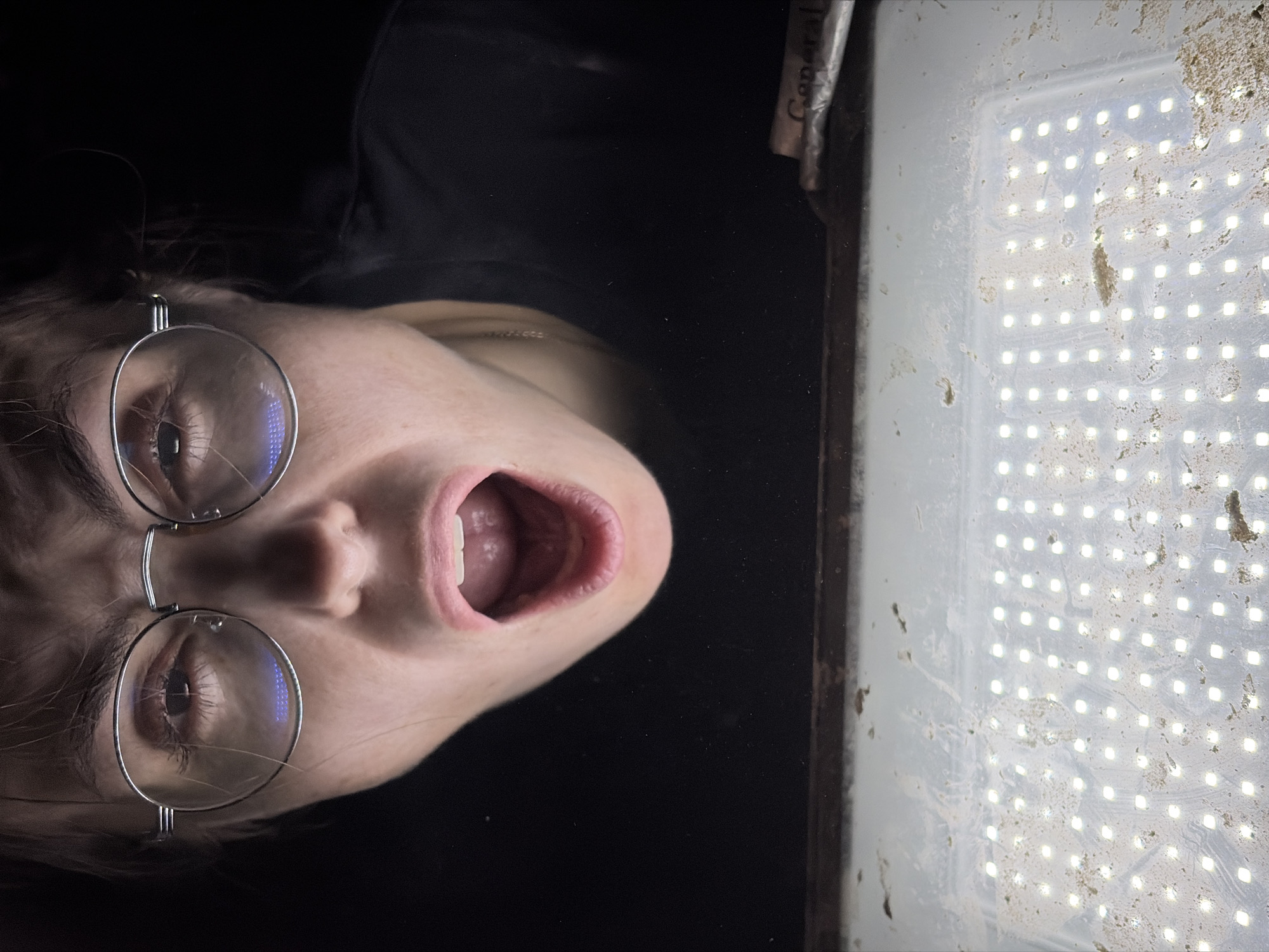 The image shows a close-up, rotated view of a person with light skin and round, metal-framed glasses, looking intently at a rectangular LED light panel. The person's face is illuminated from below by the bright light source, casting a glow on their chin and inside their open mouth. Their mouth is wide open, suggesting an expression of surprise, shock, or perhaps mid-speech. The eyes, magnified through the clear lenses of the glasses, reflect the grid pattern of the individual square LED lights. The background behind the person is entirely dark, creating a strong contrast with the brightly lit face and panel.
The LED light panel, which dominates the right side of the frame, is noticeably dirty, covered in specks of dust, dirt, and smudges, particularly evident on its white, diffused surface. Despite the grime, the grid of bright, white LEDs is clearly visible and operational. Along the top-right edge of the panel, partially obscured by shadow and the frame, the word "General" is visible on a label or object. The overall scene conveys a sense of direct interaction or observation of the light source, heightened by the person's intense expression. The location context provided is Izhevsk, Russia, though no specific visual cues for this are present in the image.