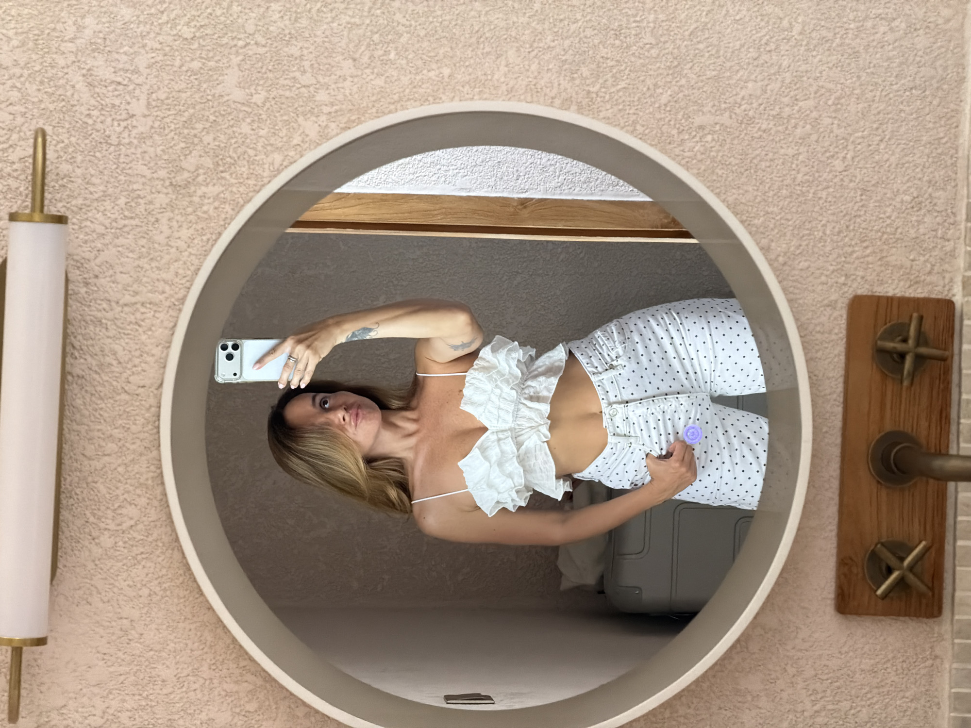 A woman is taking a selfie in a circular mirror. She is wearing a white ruffled crop top and white polka dot pants. She is standing in a room with textured walls and a light fixture above the mirror. There is a suitcase and a wooden framed object with metal fixtures visible in the reflection. The location is identified as Mengwi, Indonesia.