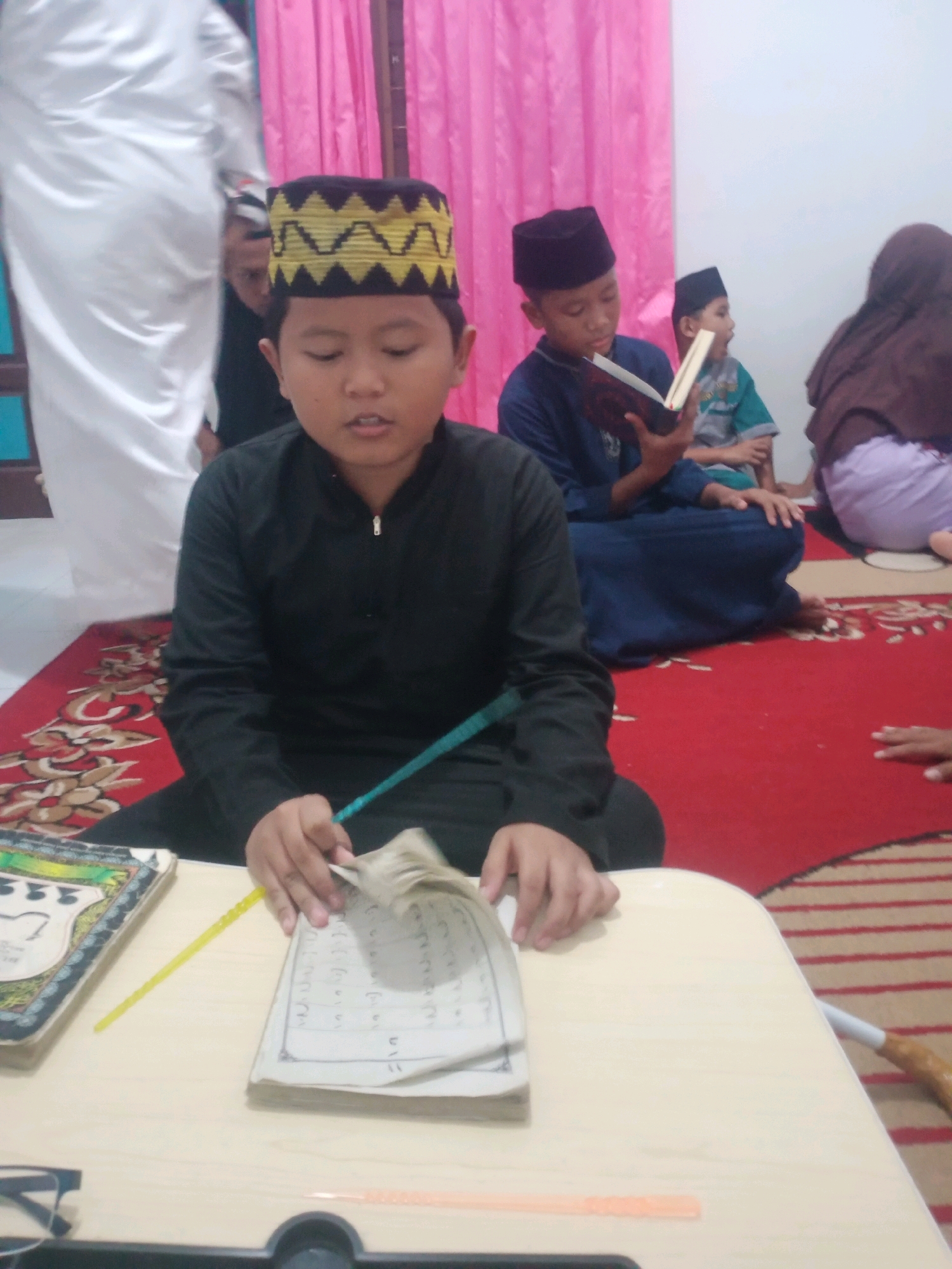 A group of children and adults, likely a family or religious study group, are gathered indoors on a red carpeted floor with a patterned rug. Pink curtains hang in the background. In the foreground, a young boy wearing a black tunic and a patterned peci (hat) is seated and looking down, holding an open book with Arabic script. Beside him, another child in a blue tunic is reading a book. To the right, an adult in a brown hijab is visible, and further back, a person in white traditional attire is standing. The scene appears to be a casual gathering, possibly for religious education or prayer, in a residential setting within Bandar Lampung, Indonesia.