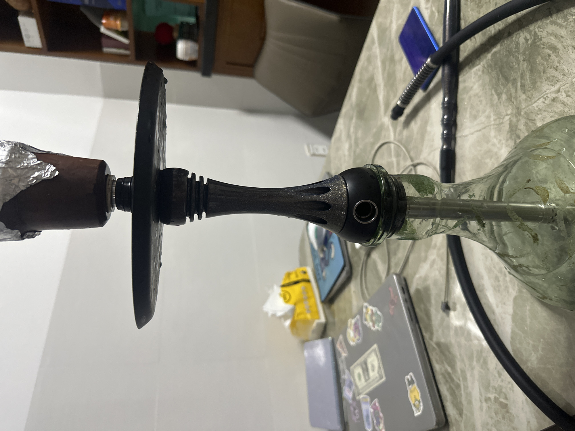 The image shows a hookah on a marble-like surface. The hookah has a silver-covered bowl, a dark stem, and a clear glass base partially filled with liquid. A black hose is connected to the base. Scattered around the hookah are a blue card, a box of tissues, a laptop with stickers, and other miscellaneous items. The background includes a cabinet and a chair. The location is Bac Nha Trang Ward, Vietnam.