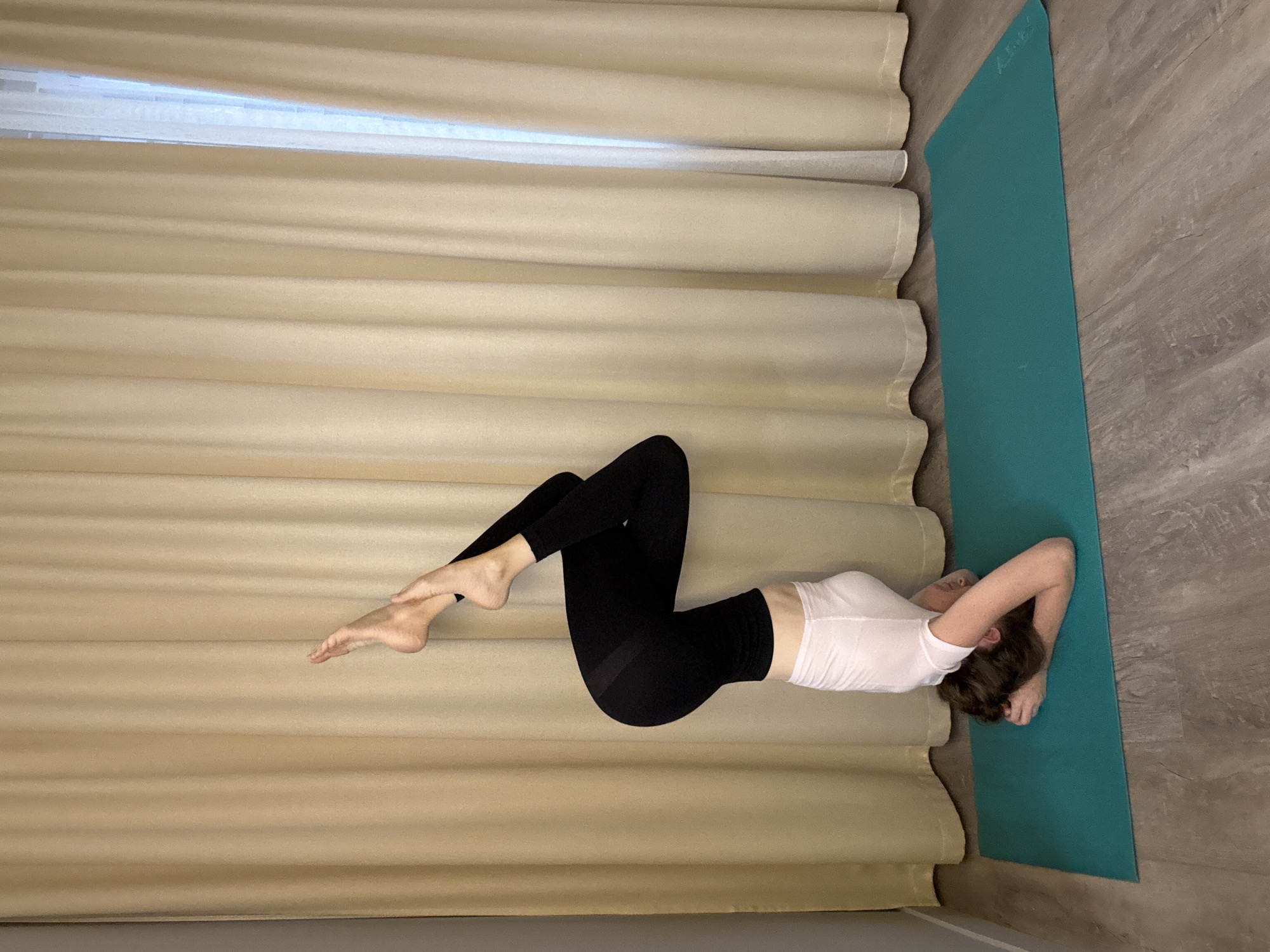 A person is performing a yoga pose, specifically a headstand variation, on a teal yoga mat. The person is upside down, with their head and forearms on the mat. Their legs are bent and crossed, with one foot extended upwards. They are wearing a white t-shirt and black leggings. The background consists of beige curtains and a wooden floor. The scene appears to be indoors, possibly a home or yoga studio. No specific location in Dolgoprudny, Russia can be determined from the image.