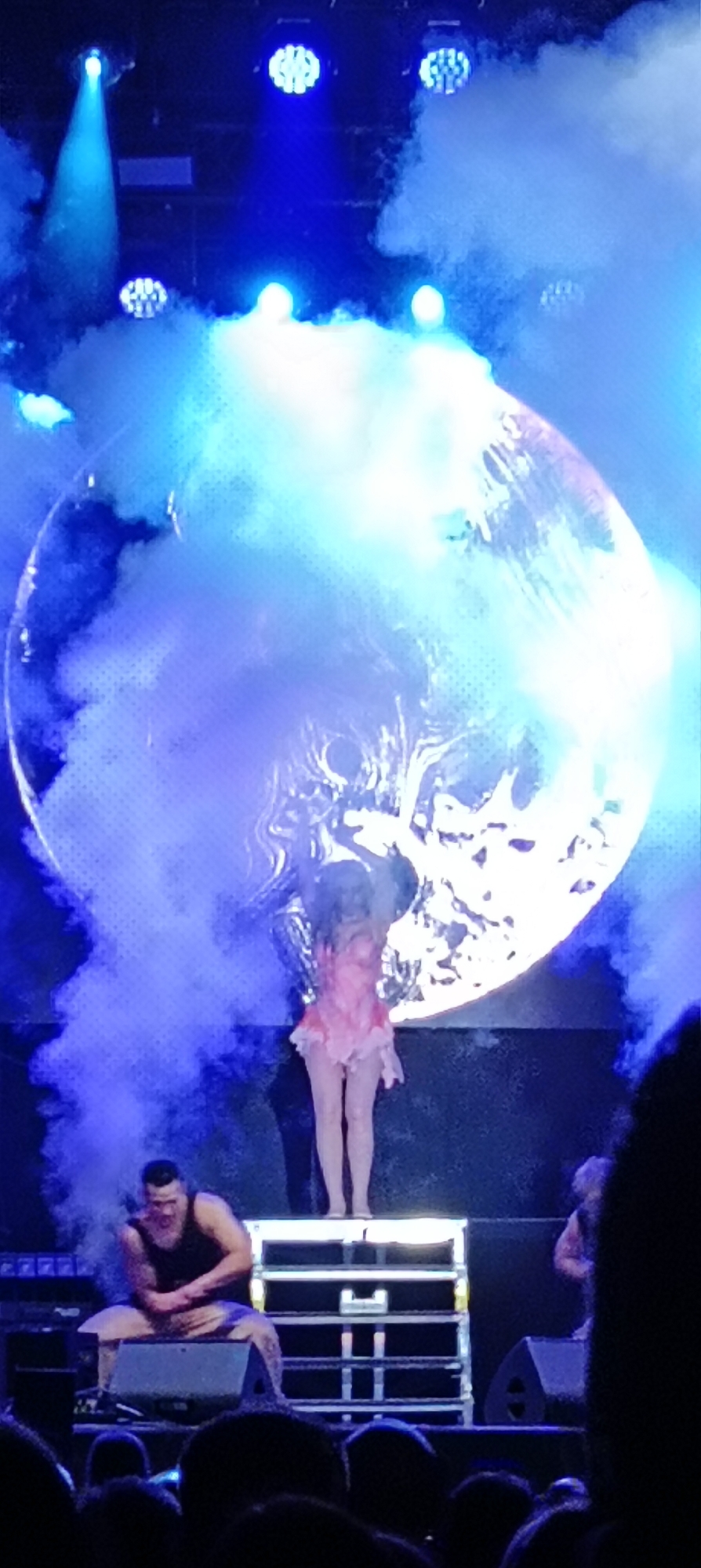A view from an audience at a concert scene in Cerdanyola del Vallès, Spain. A person stands center stage in a pink outfit in front of a large illuminated backdrop. A man sits on the stage to the left of her. Smoke from the stage partially obscures the view. The scene is lit with blue stage lights. The foreground is filled with the heads of other audience members.