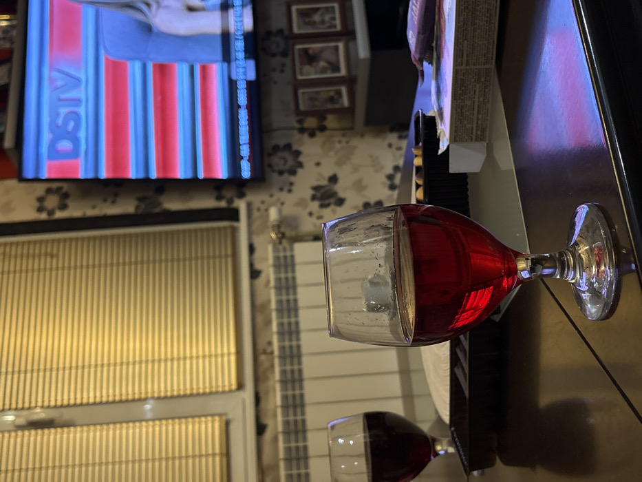 The image depicts an indoor residential setting in Cherven Bryag, Bulgaria, characterized by dim ambient lighting. In the foreground, positioned on a dark, reflective table surface, is a clear, stemmed glass containing a dark red liquid, likely wine. Condensation or small water droplets are visible on the exterior surface of this glass. To the left and slightly behind, a second, similarly filled stemmed glass is partially visible on the same table.
Behind the foreground glass, a black rectangular tray or container is present, alongside a partially visible white object and a cardboard box with visible text on its side.
The background features a window with closed light-colored horizontal blinds on the left. A white central heating radiator is mounted below the window. On the right, a flat-screen television is displaying content with the "DSTV" logo, characterized by horizontal red and blue graphical bands, and a partial view of a person. The television is mounted on a wall covered with patterned wallpaper, dark in tone with lighter floral or abstract motifs. Below the television, a dark piece of furniture holds three framed pictures arranged horizontally. No individuals are present within the visible frame. The overall scene suggests a prepared environment for relaxation or entertainment.