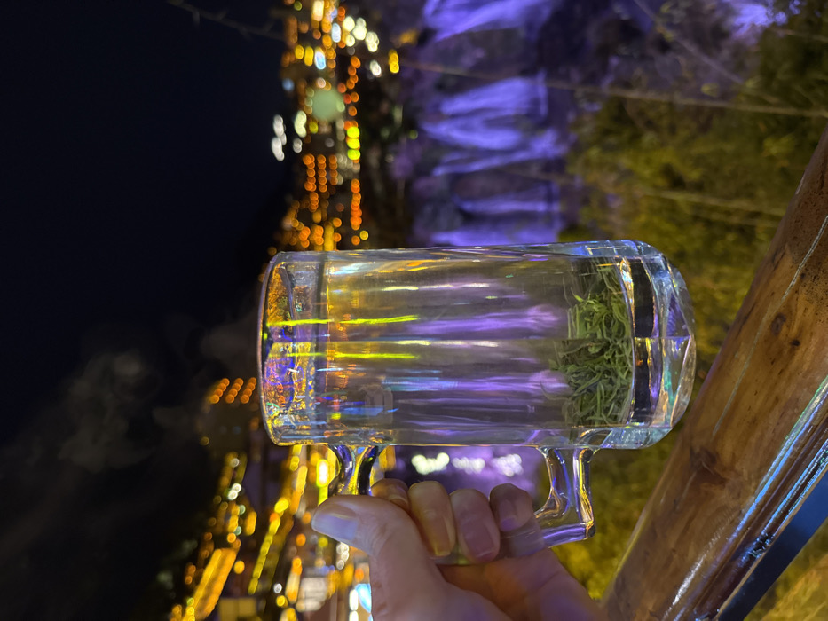 A hand holds a clear glass mug containing steaming hot water and green tea leaves. The mug's surface reflects vertical yellow and purple light streaks from the environment. In the background, the night scene of Furong Ancient Town, China, unfolds. Blurred city lights, predominantly orange and yellow, illuminate a distant hillside to the left. To the right, multiple waterfalls are visible, illuminated with a purple light, descending into blurred green vegetation below. The sky overhead is dark, indicating nighttime. A wooden railing runs diagonally across the lower left foreground.
