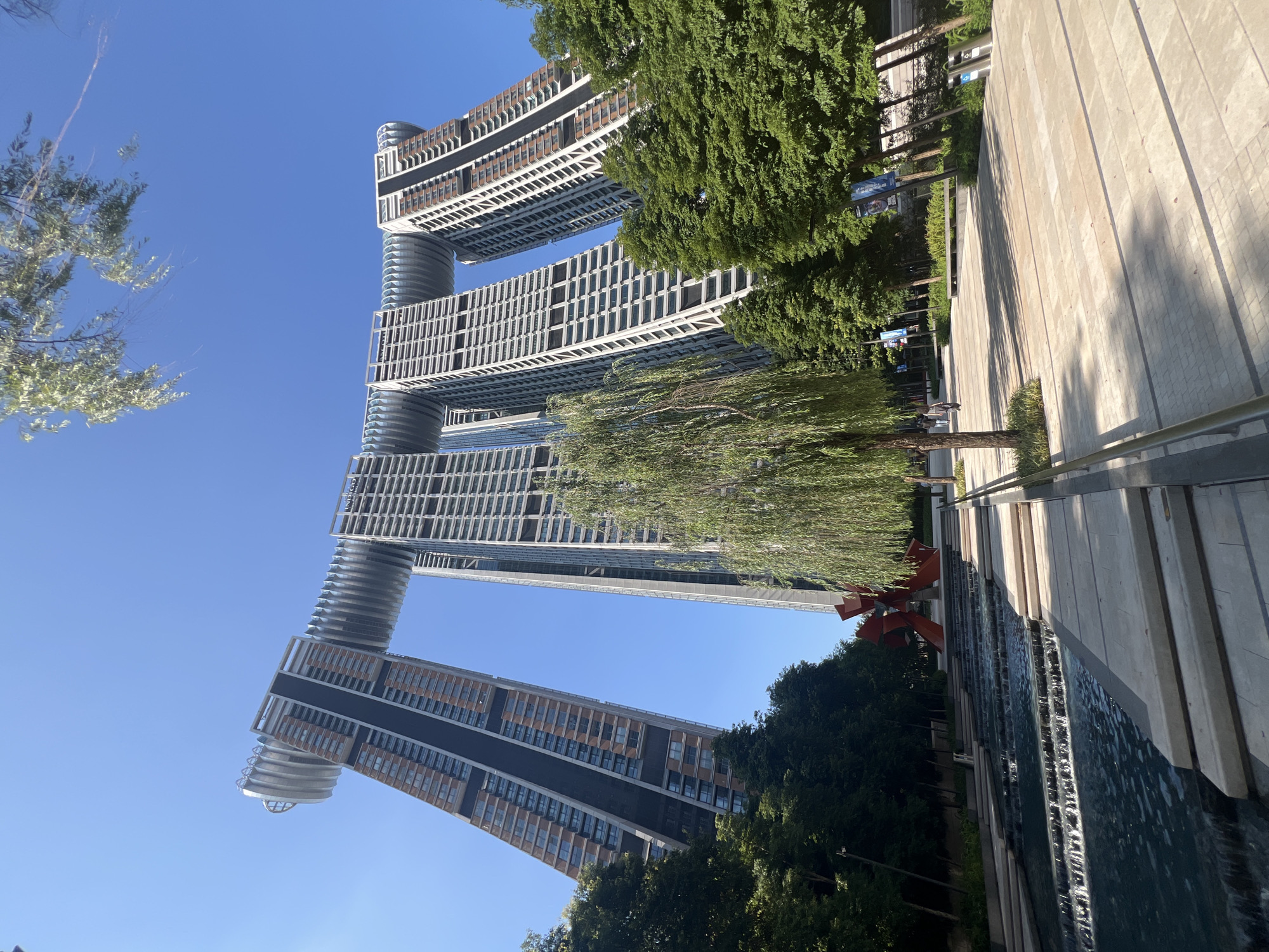 The image shows the landmark "Crystal" building complex in the Yuzhong District of Chongqing, China. The complex features four residential towers connected by a horizontal sky bridge, forming a distinctive archway shape. The towers are clad in a mixture of glass and dark paneling, with a ribbed, metallic structure forming the sky bridge. In the foreground, a tiered water feature cascades down steps, with a paved walkway and lush green trees flanking it. A contemporary red sculpture is visible in the background, nestled amongst the trees. The sky is a clear, bright blue. Several individuals are walking on the paved pathway in the middle ground.