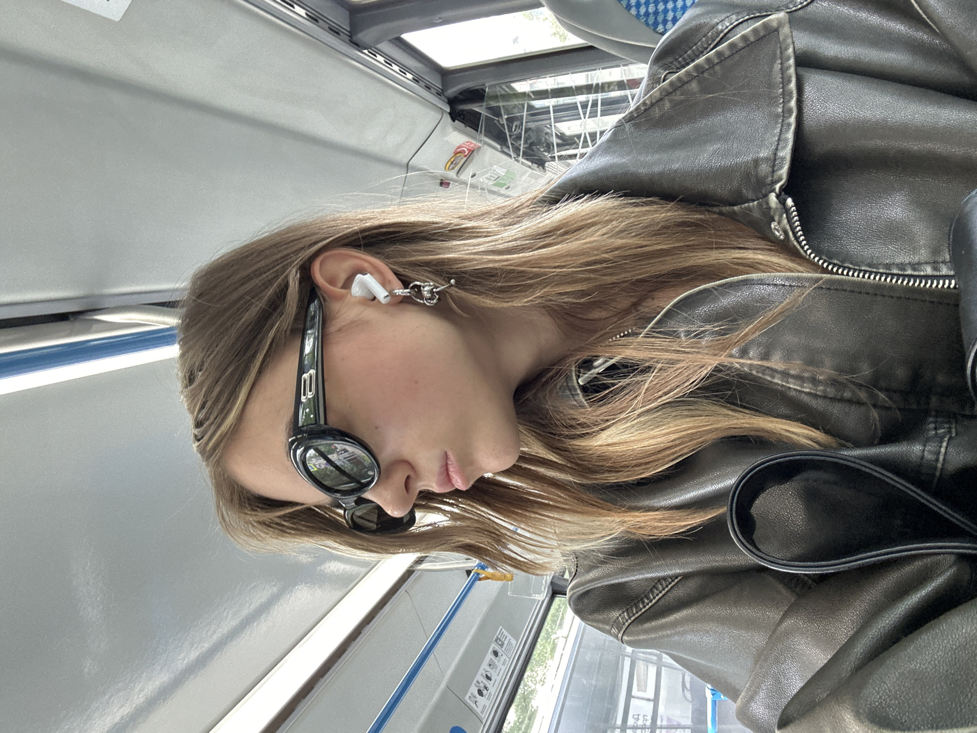 A young woman with long blonde hair is seated on a bus, looking out the window to her left. She is wearing black oval-shaped sunglasses, white wireless earbuds with a dangling silver earring, and a dark brown leather jacket. The interior of the bus is visible, with blue handrails and windows showing a blurred outdoor scene.