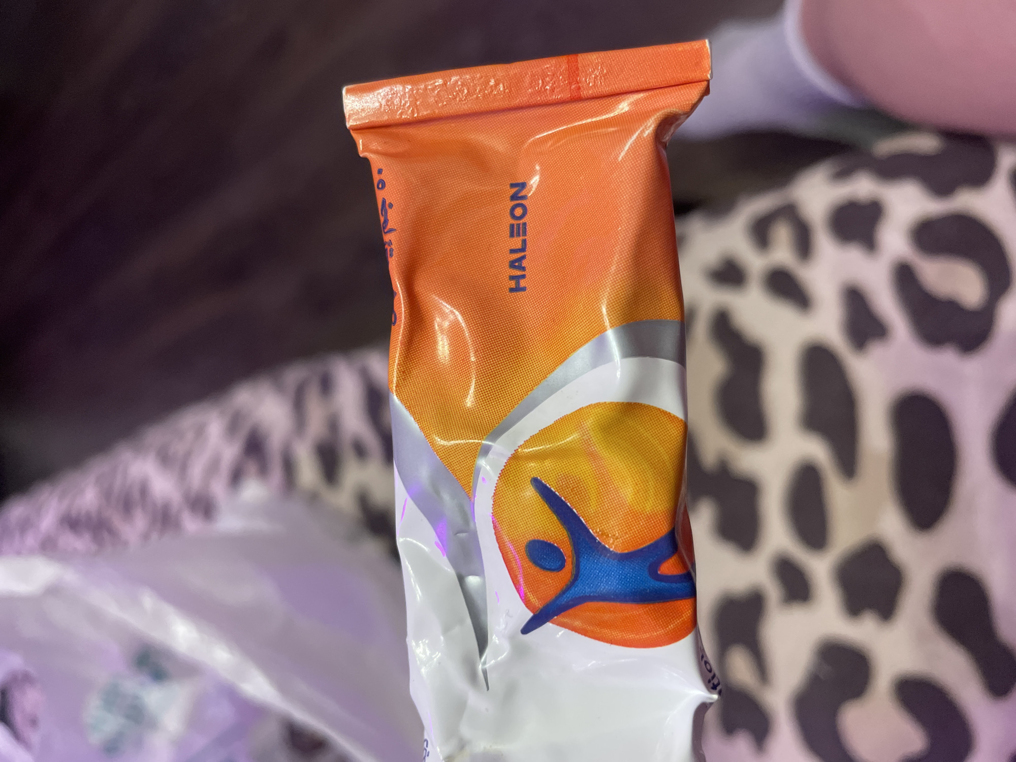 The image shows a close-up of a product packaging, which is primarily orange and white with blue and yellow accents. A blue human-like figure with outstretched arms is depicted on the white portion, set against a yellow and orange circular background. The word "HALEON" is clearly visible in blue text on the orange section of the packaging. The product is held at an angle, with the right edge of the packaging folded over, revealing a textured orange border. In the background, a person's legs are visible, wearing white socks and a light-colored garment with a leopard-like print. A portion of a white plastic bag is also visible in the upper left corner. The scene appears to be indoors, with the lighting suggesting it's not daytime. There is no indication of Vaganovo, Russia, or any specific city in the image.