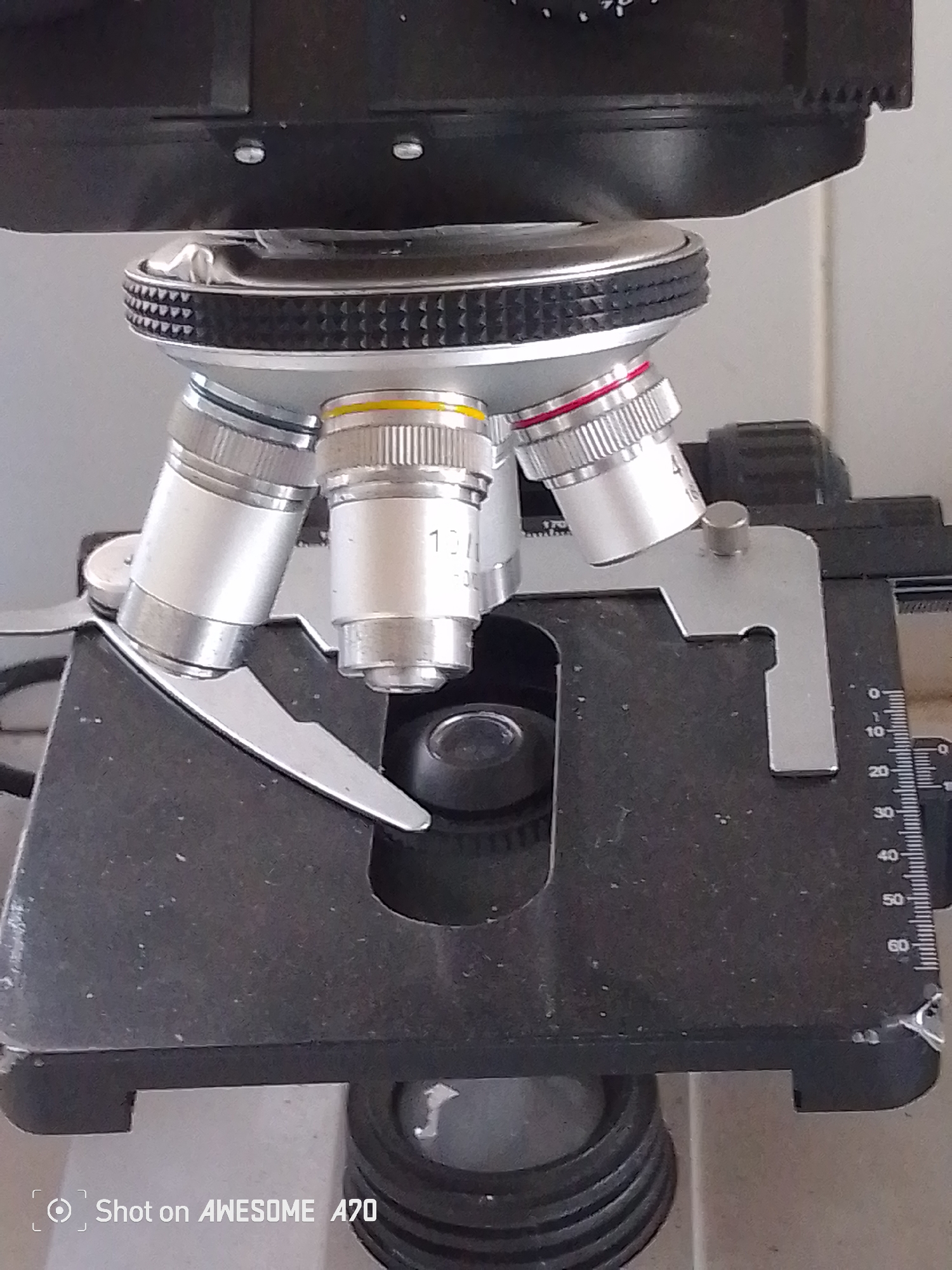 The image is a close-up, low-angle shot of the objective lens turret and stage of a microscope. Three objective lenses are visible, two of which have magnification labels indicating "10X" and "40X" respectively. A portion of the stage with a ruler marking appears on the right side of the image. The microscope is black with silver metallic accents on the lenses and turret. The background is a plain, light-colored wall. The bottom of the image contains a watermark that reads "Shot on AWESOME A70". There are no people or any indication of a specific location within the city of Sukabumi, Indonesia.