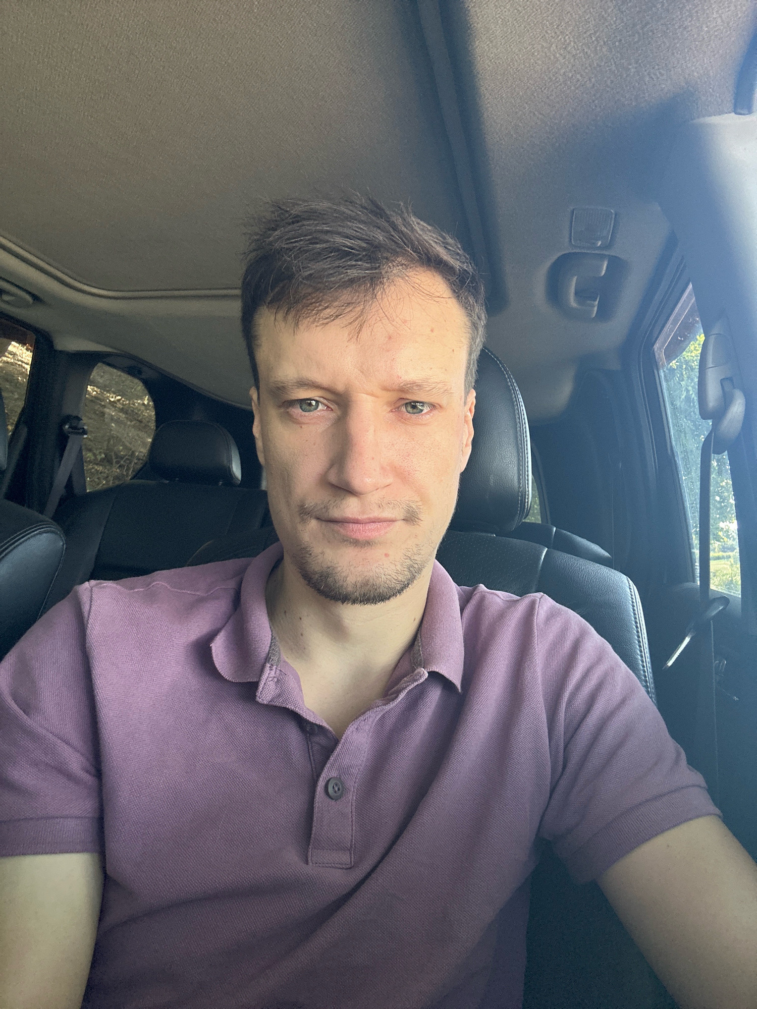 A man with short, brown hair and a light beard is seated in the driver's seat of a car, looking at the camera. He is wearing a purple polo shirt. The interior of the car is visible, including black leather seats, a sunroof, and the window on the right side of the car. Outside the window, trees are visible, suggesting a rural or suburban setting, but no specific landmarks can be identified to pinpoint the location in Makiv, Ukraine. The lighting suggests it is daytime, with sunlight entering the car.