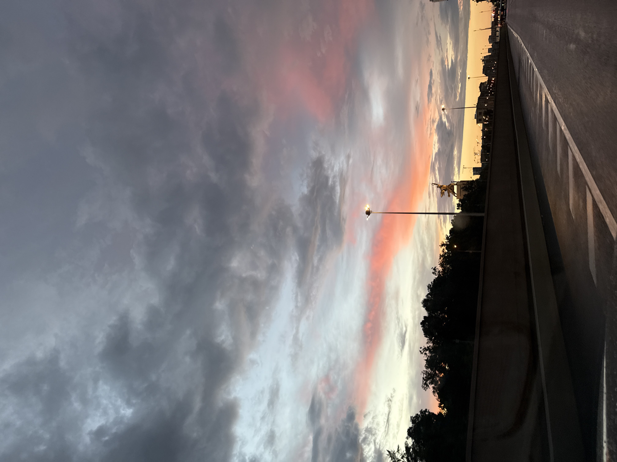 This media file depicts a city skyline at dusk, with a focus on the sky as it transitions from day to night. The sky is filled with a dramatic mix of clouds, with dark gray clouds on the left side of the frame gradually giving way to streaks of pink and orange as the sun sets on the horizon. The overall mood is one of twilight and impending night.
The scene includes a road with white markings on the right, suggesting a highway or main thoroughfare. A dark concrete barrier separates the road from a strip of trees, which are silhouetted against the colorful sky. Beyond the trees, a city skyline is visible, with buildings and streetlights creating a man-made element against the natural beauty of the sunset. A street lamp with a bright light is prominent in the mid-ground, illuminating the immediate area. In the distance, a statue or monument is visible, adding a point of interest to the urban landscape.
The time of day is clearly dusk, as indicated by the sunset colors in the sky and the streetlights that are beginning to illuminate. The weather appears to be partly cloudy, with the mixture of dark and lighter clouds contributing to the dramatic effect. The location is identified as Moscow, Russia, which suggests the skyline and any visible structures may be characteristic of that city. There is no visible text in the media file.