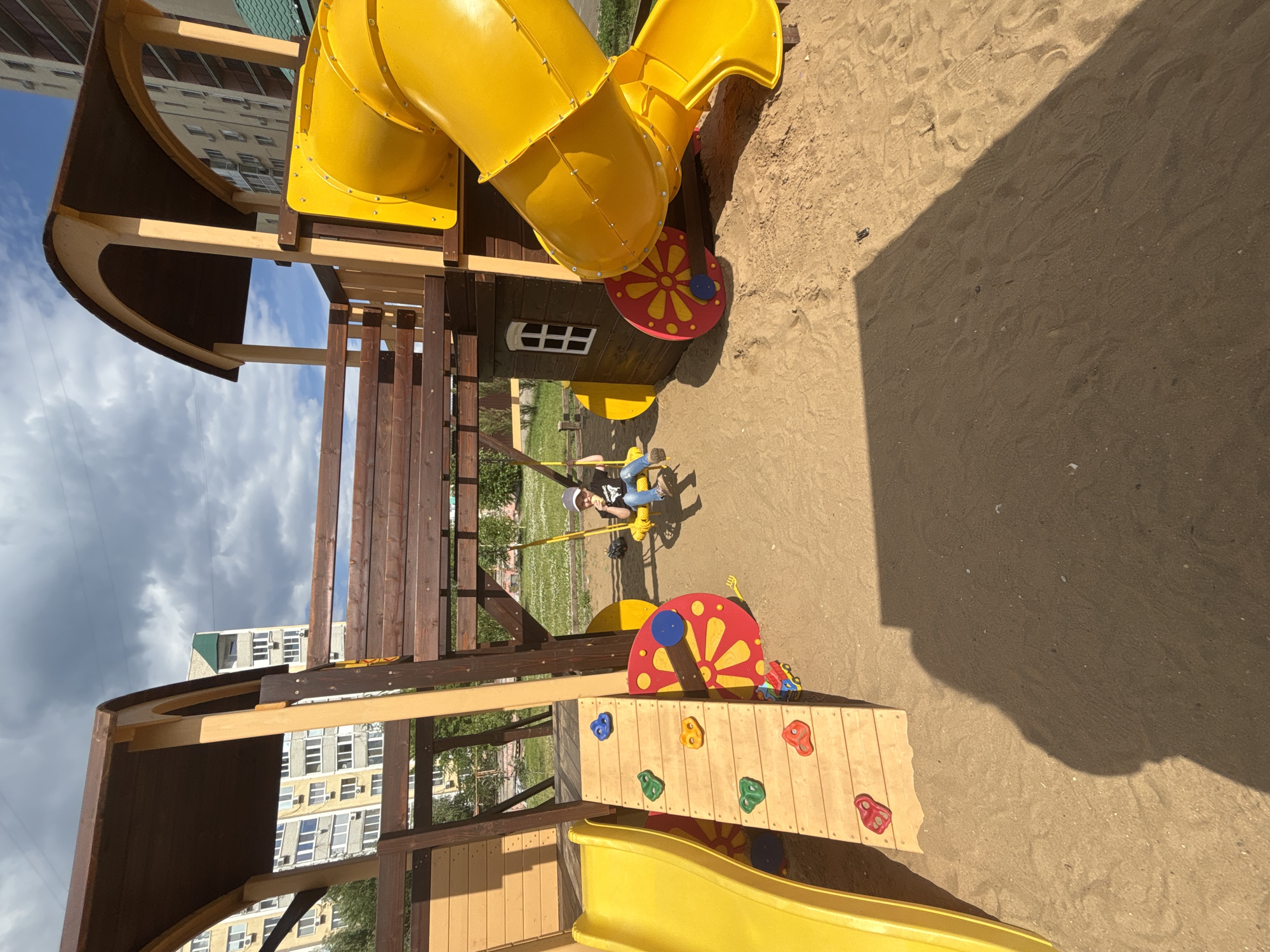 The image depicts a children's playground in Saratov, Russia, on a cloudy day. The playground features a large, multi-level wooden structure with a yellow slide, a climbing wall with colorful handholds, and a yellow tube slide. A young child, wearing a dark t-shirt, blue jeans, and a grey cap, is seated on a yellow swing, holding a white object, possibly food. The playground is situated on a sand-covered surface, with grassy areas and residential buildings visible in the background. The shadow of the photographer is cast across the sand in the foreground.
