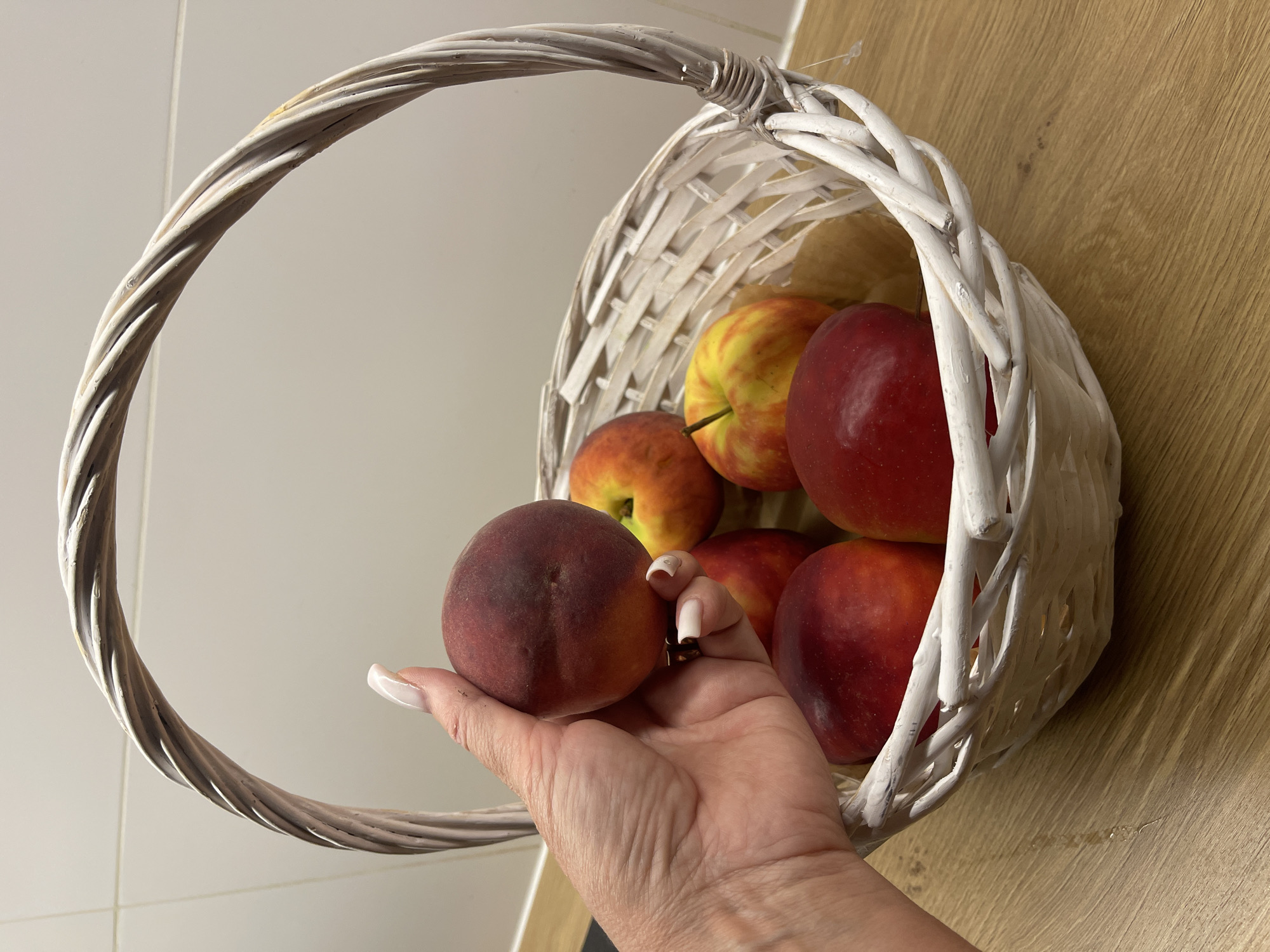 A person's hand, with painted fingernails, holds a dark red nectarine above a white wicker basket filled with several nectarines and apples. The basket sits on a wooden surface, likely a countertop, with a white tiled wall in the background. The scene appears to be indoors, possibly a kitchen, but there is no specific information to determine the location in Wrocław, Poland. The lighting suggests artificial light is present.