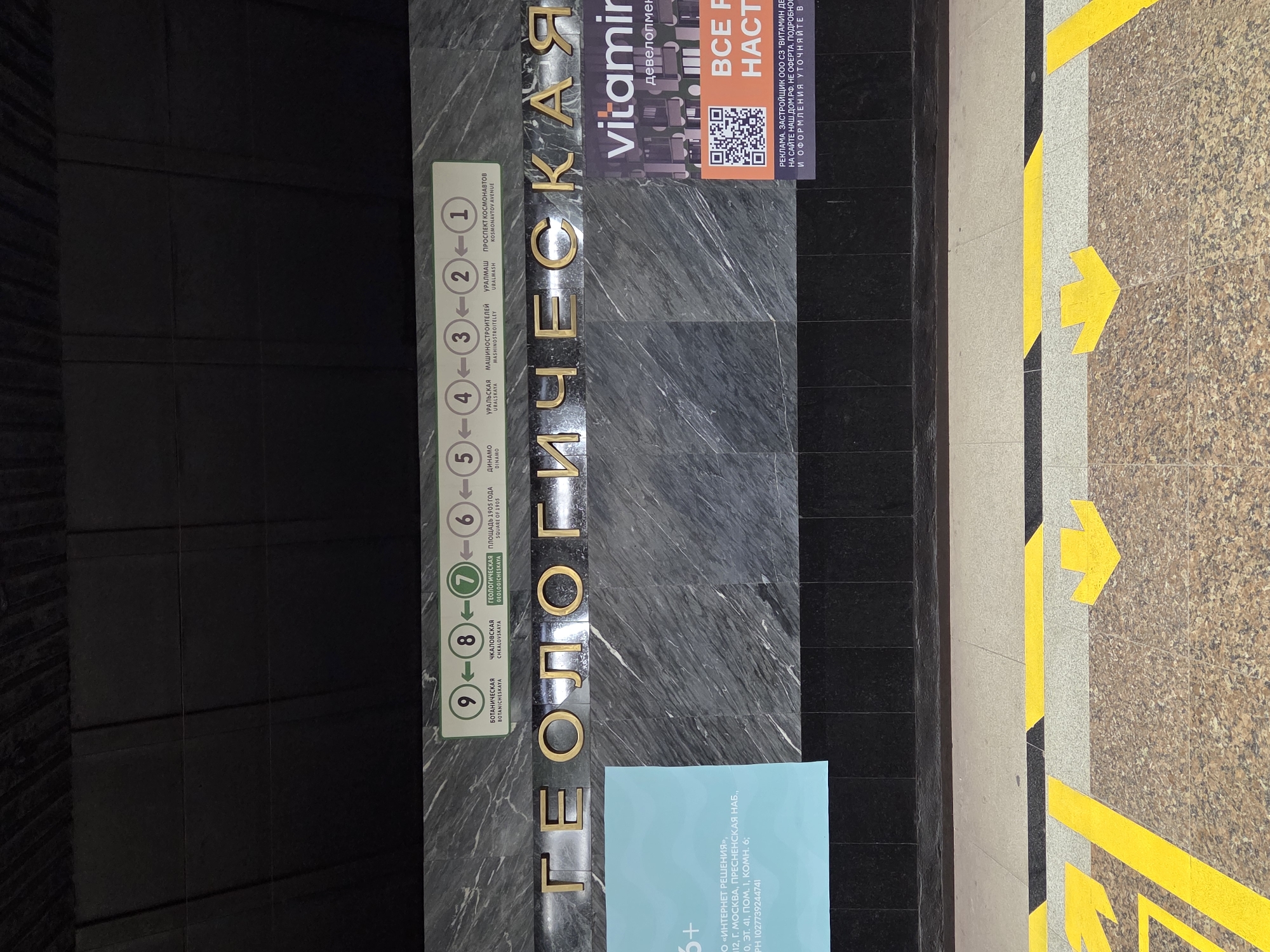 The image shows the interior of a metro station in Yekaterinburg, Russia. The station name, "GEOLOGICHESKAYA" (ГЕОЛОГИЧЕСКАЯ), is displayed in large gold letters across a dark grey, marble-patterned wall. Above the station name, a line of green circles with white numbers and arrows indicates the metro line and adjacent stations: 9 (BOTANICHESKAYA), 8 (CHKALOVSKAYA), 7 (GEOLOGICHESKAYA), 6 (PLOSHAD 1905 GODA), 5 (DINAMO), 4 (URALSKAYA), 3 (MASHINOSTROITELEY), 2 (URALMASH), and 1 (PROSPEKT KOSMONAVTOV). To the left of the station name, a light blue poster with white text is partially visible. To the right, a smaller advertisement with the word "vitamin" in white lettering is displayed on a purple background, along with a QR code and the text "ВСЕ РЕШЕНИЯ НАСТОЯЩЕ" (All solutions are present). The foreground of the image shows the station platform with yellow arrows painted on the floor, indicating directions of movement. The station appears to be clean and modern, with no people visible in the frame.