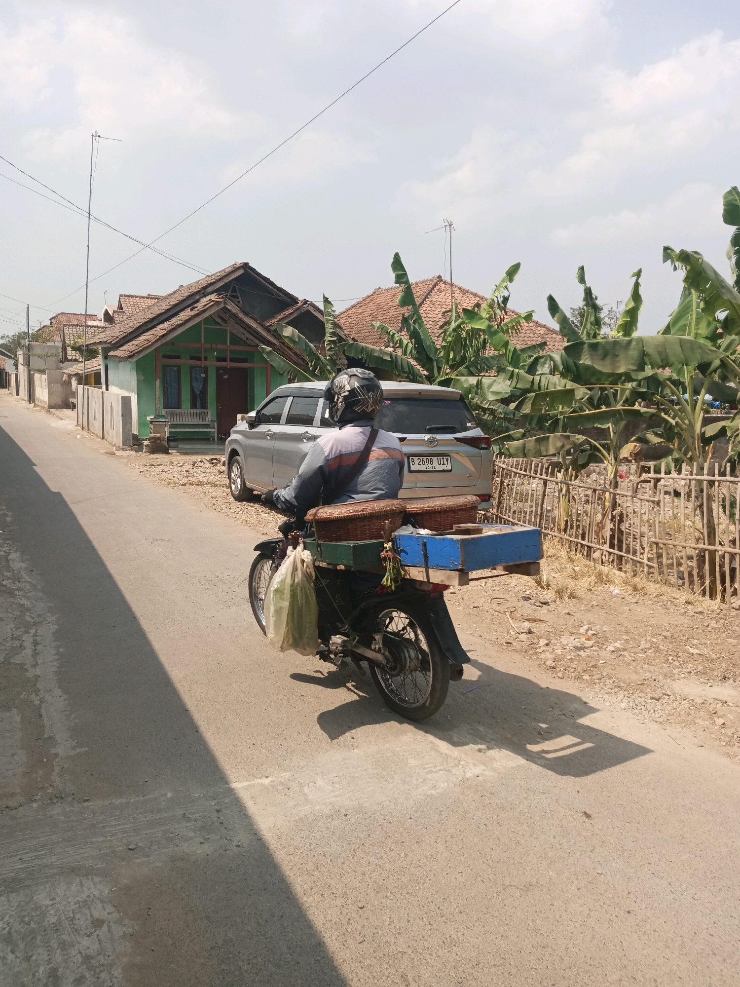 In Subang, Indonesia, a person rides a motorcycle on a paved road. The motorcycle is dark-colored with a basket attached to the rear, holding a blue box and a woven basket. A light-colored plastic bag hangs from the motorcycle. The rider wears a helmet and a blue and orange jacket. A silver car with license plate B 2698 UIY is parked on the side of the road. In the background, there are several houses with terracotta roofs and green walls. Banana trees are visible, and a bamboo fence borders the property. The sky is partly cloudy.