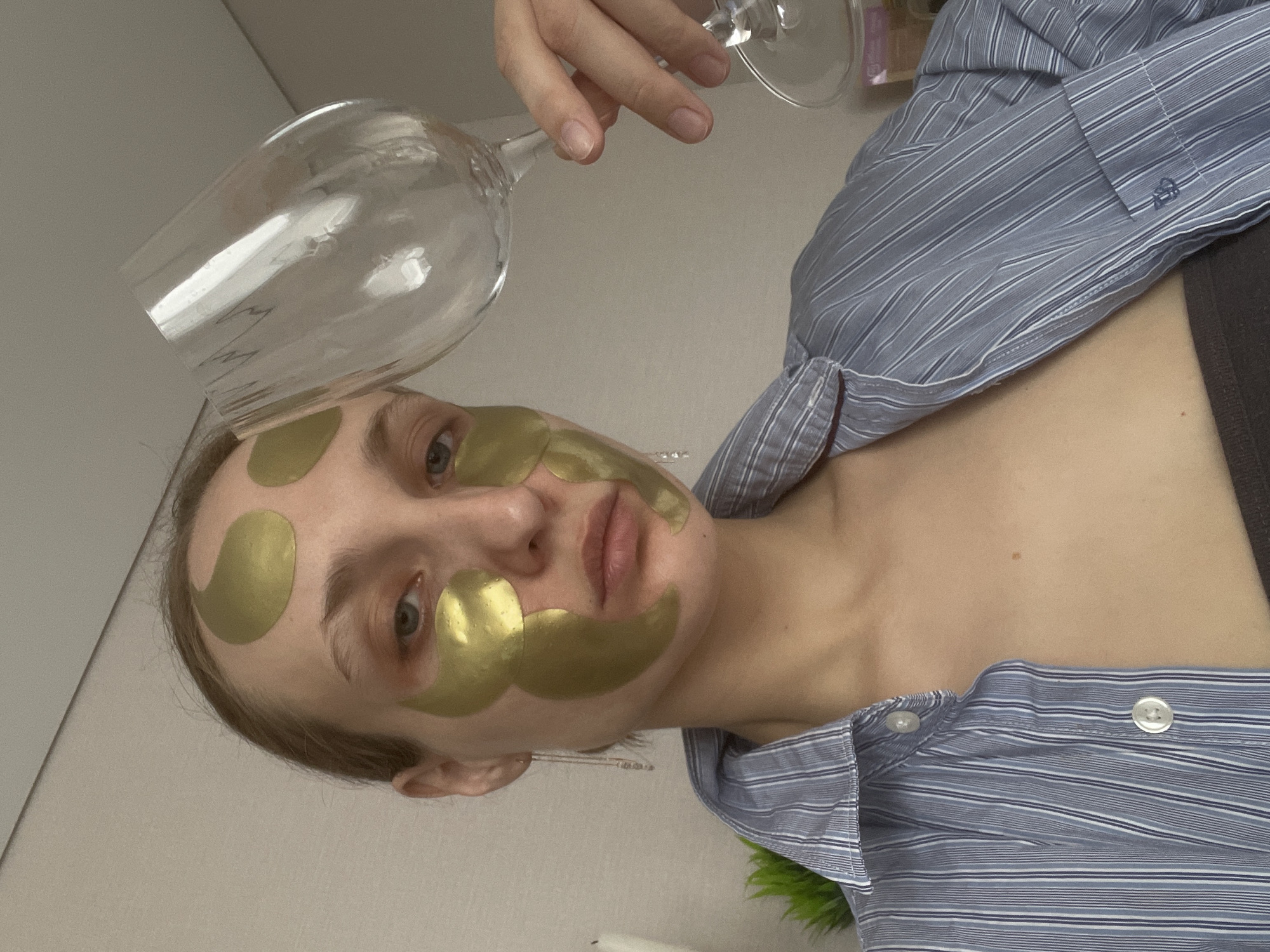 A person is holding a wine glass over their face, which has golden patches on the forehead and under the eyes. The person is wearing a striped blue shirt. The scene appears to be indoors with a plain wall in the background. There is no evidence to suggest that the picture was taken in Подшивалово, Russia.