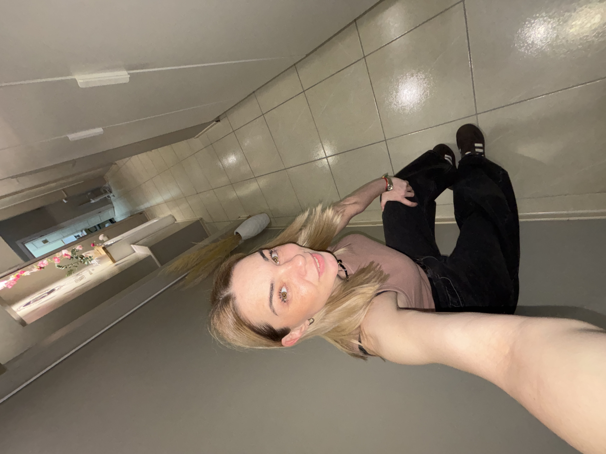 A person with blonde hair, wearing black pants and brown shoes with white stripes, is lying on a tiled floor in a hallway. The hallway has a long, narrow opening that looks down into a lower level. The lower level contains a floral arrangement and other furnishings. The location is Unknown, Unknown.