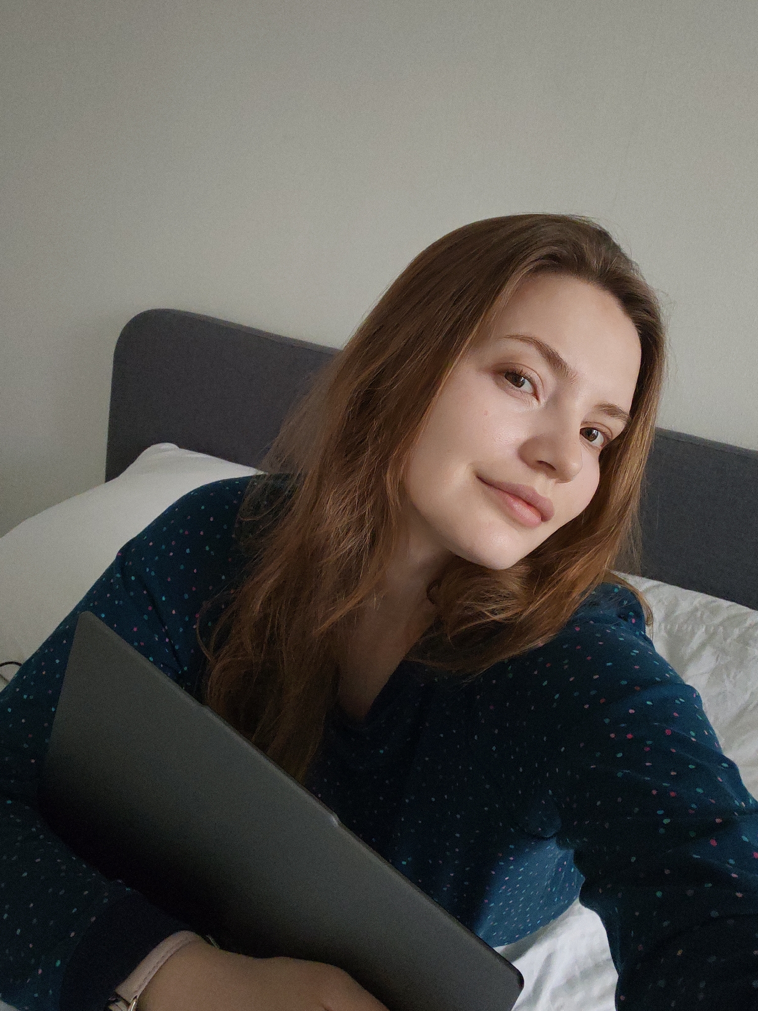 A young woman is captured in a close-up, high-angle shot, appearing to take a selfie or engage in a video call from a bed. She has light skin, long, wavy, reddish-brown hair, and is looking directly at the camera with a gentle, soft smile, conveying a pleasant and relaxed demeanor.
She is wearing a dark teal or navy long-sleeved top patterned with small, colorful polka dots in pink, blue, and light green hues. A light pink or rose gold watch is visible on her left wrist. Her hair frames her face and extends over her shoulders and chest. Her facial features include clear skin, natural-looking eyebrows, and subtly tinted lips.
The woman is holding a dark-colored laptop or tablet, which is either closed or in a protective case, resting against her body. She is positioned in a bed with white bedding, including a pillow and a duvet or sheet. A dark gray padded headboard is visible behind her. The background consists of a plain, light-colored wall, indicating an indoor, likely bedroom, setting. The lighting suggests daytime. The image has a slight grainy texture. No visible text is present.