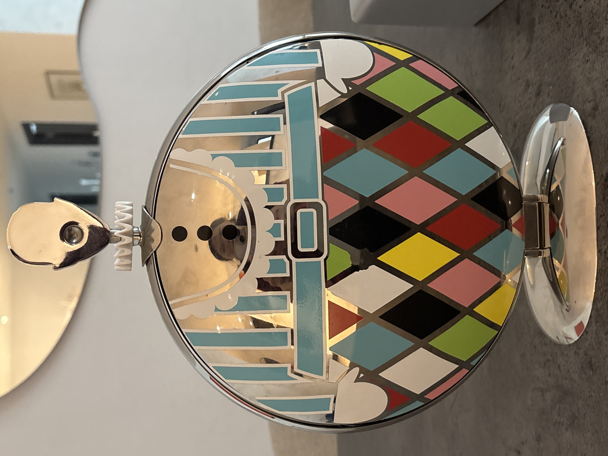 A close-up, eye-level shot shows a circular, reflective object designed to resemble a clown's face. The object is silver and features a stylized headpiece at the top, a ruffled collar below it, and a colorful diamond pattern on its lower half. The diamond pattern includes various colors such as blue, white, pink, red, green, yellow, and black. The object sits on a mirrored base. In the reflection on the object's surface, a person's face is visible. The background appears to be an indoor environment with a wall and a table. The location is Dubai, United Arab Emirates.