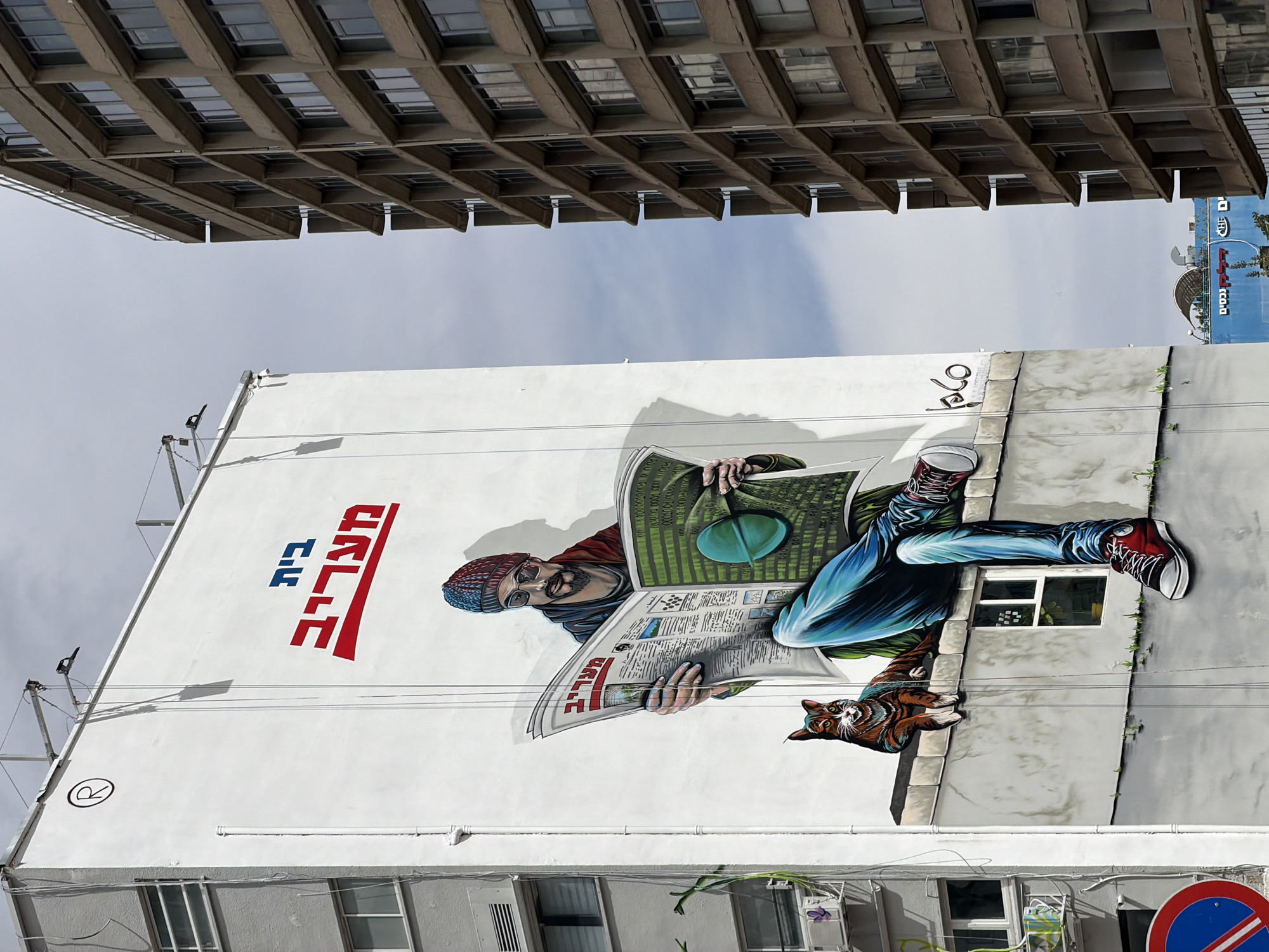The image shows a mural on the side of a building in Tel Aviv, Israel. The mural depicts a man wearing a beanie and glasses, reading a newspaper while reclining on what appears to be a windowsill. A cat lies nearby. The Hebrew words for "Haaretz" (a newspaper) are visible on the mural, along with the artist's signature "rhaj". A modern, brutalist-style building looms above. A glimpse of the Mediterranean Sea and other signs in Hebrew are visible in the distance.
