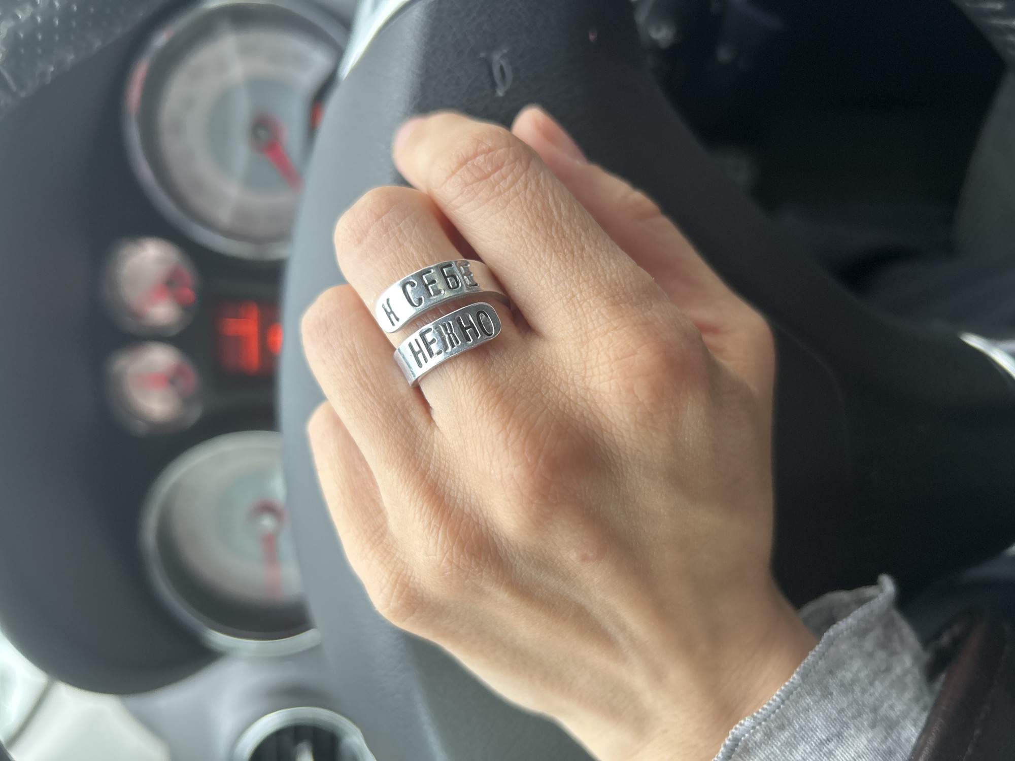 A close-up image shows a person's right hand resting on a car's steering wheel. The hand, appearing slender, wears a distinctive silver-colored, two-band wrap-around ring on the ring finger. The top band of the ring features the Cyrillic text "К СЕБЕ" (K SEBE), and the bottom band displays "НЕЖНО" (NEZHNO), which together translate to "To myself gently" or "Be gentle with yourself."
The background is a blurred interior of a car, with elements of the dashboard visible, including out-of-focus gauges like a speedometer and tachometer, both showing red needles. A grey garment, possibly a sleeve or collar, is partially visible on the left side of the frame. The lighting suggests natural daylight. The image focuses on the personal detail of the hand and ring within the context of a car. Given the Russian text on the ring, the provided location context of Moscow, Russia, is consistent. The activity appears to be the hand resting on the steering wheel, indicating the person is either driving or a passenger in the vehicle.