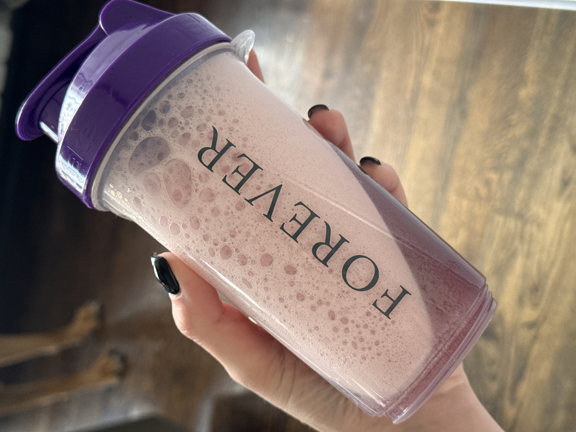 A Caucasian hand with black nail polish holds a clear plastic shaker cup filled with a frothy pink liquid. The cup has a purple lid and is printed with the word "FOREVER" in black, vertically oriented text. The background shows a wooden floor and partially visible dog legs, suggesting an interior location. The image was not taken in Sofia, Bulgaria.