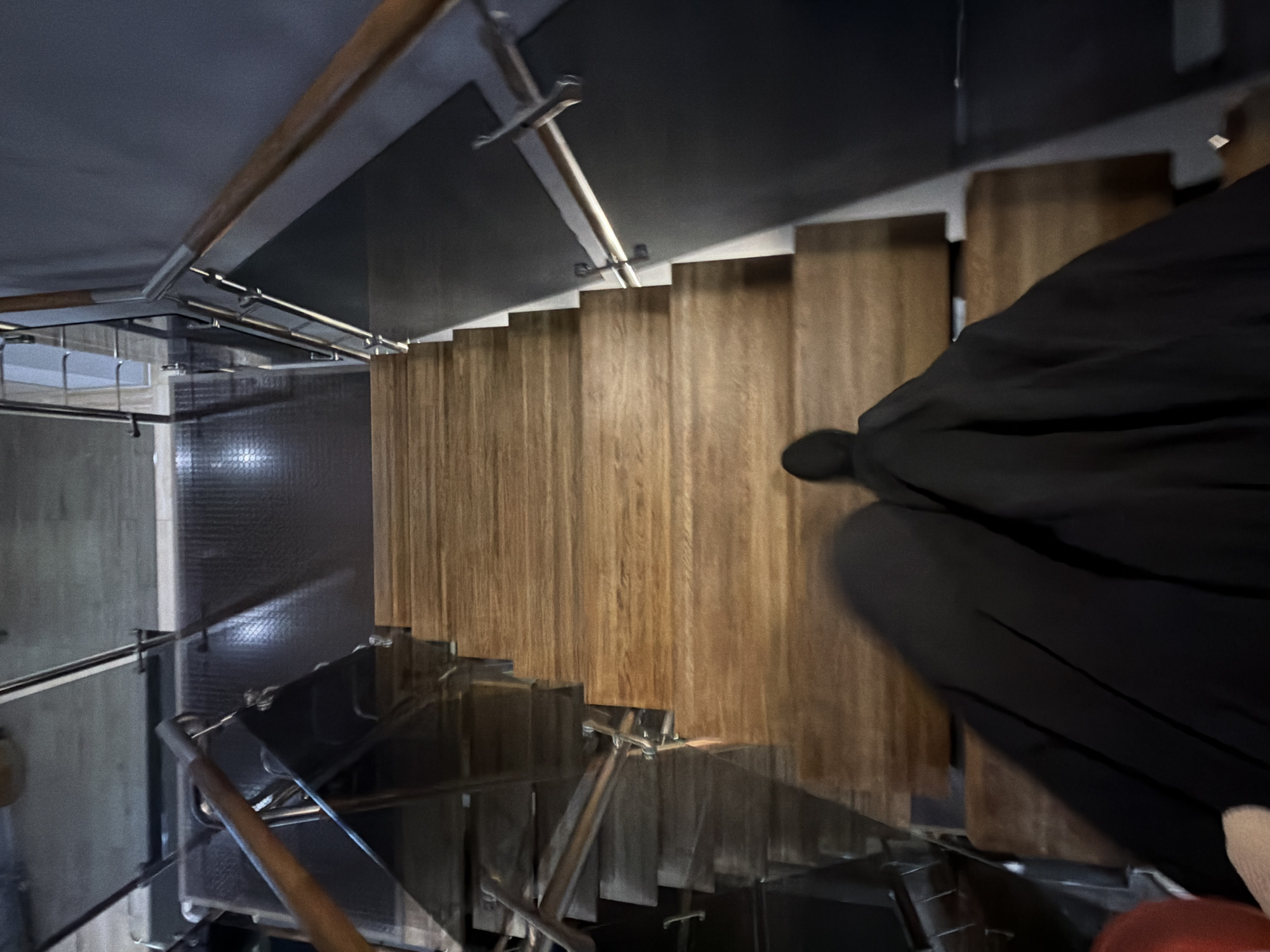 A high-angle shot depicts a staircase with wooden steps enclosed by metal and glass railings, with a person dressed in black descending. The stairwell has dark ceilings and walls, with some reflective surfaces. The overall setting suggests a modern interior design. The location in the city of Saint Petersburg, Russia cannot be determined based solely on this image.