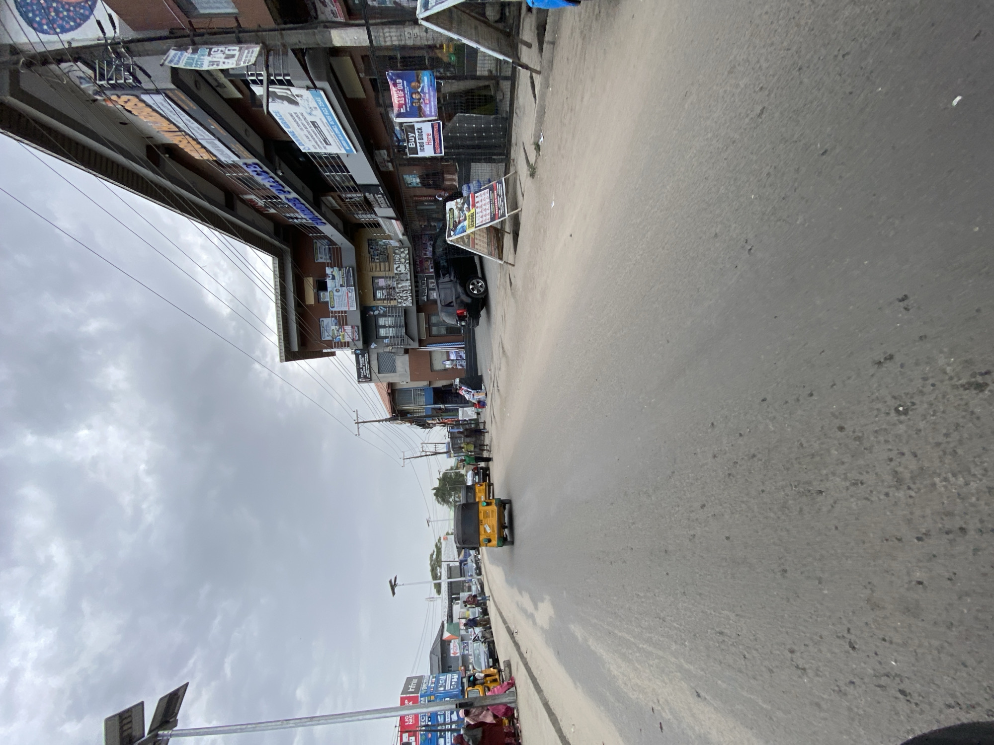 The image shows a street view in Ado, Nigeria. The road is paved and takes up a significant portion of the frame. Buildings line the left side of the street, with multiple stories and various advertisements and signs. Power lines are visible above the buildings. Several people are present on the street, as well as yellow auto rickshaws. The sky is overcast with a cloudy appearance.