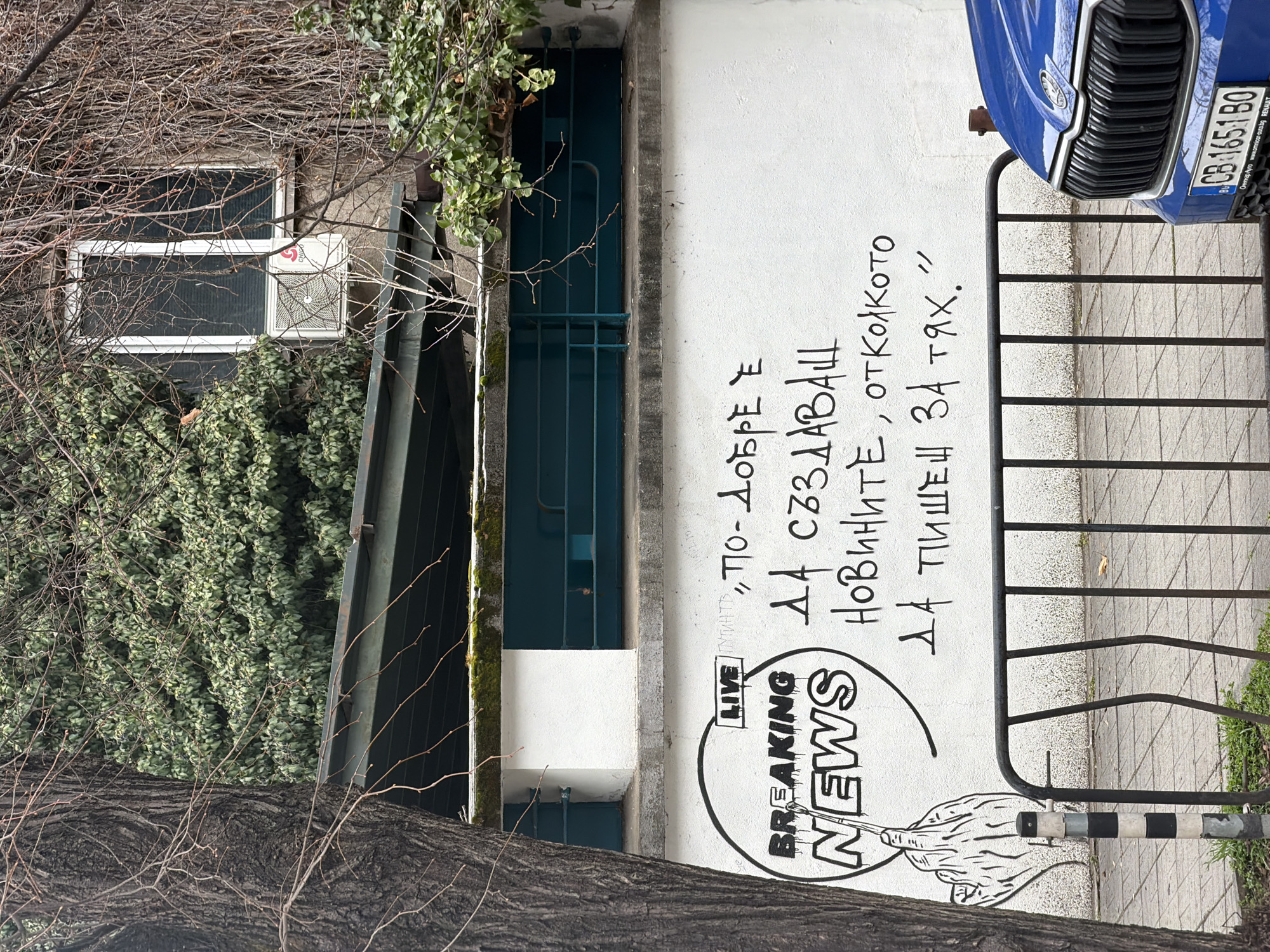 This image captures a street scene, likely in Sofia, Bulgaria, indicated by the car's license plate. A light-colored wall is the primary backdrop, featuring black graffiti art and Cyrillic text, including the phrase "Breaking News" and a drawing of a hand with an extended middle finger. The text roughly translates to "It's better to create news than to write about them." A blue Skoda car is parked beside the wall, and a black metal fence separates the wall from the sidewalk. A tree trunk with bare branches is visible in the foreground, partially obscuring the scene. The environment also includes an air conditioner unit mounted near a window, adding to the urban feel of the location.