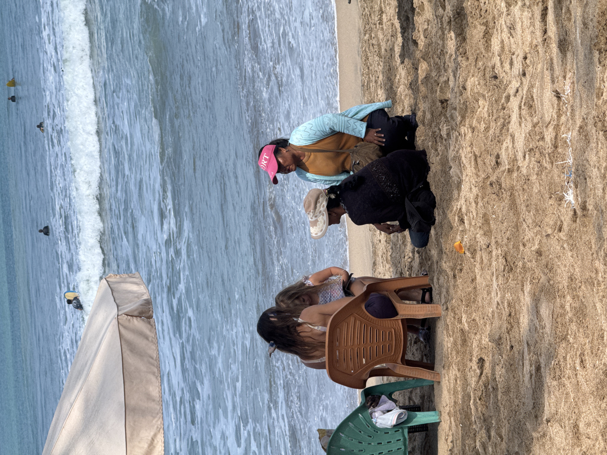 The image depicts a beach scene in Kuta, Indonesia, with the ocean and sand as the primary environment. In the foreground, three women and a young girl are gathered on the sand. Two women are seated on chairs, one in a brown plastic chair and another in a green plastic chair, partially visible. A young girl stands next to the woman in the brown chair, appearing to interact with her. Two other women are seated on the sand in front of the chairs. One woman is wearing a black top and a hat, and the other is wearing a light blue cardigan over an orange top and a pink visor. The ocean in the background shows gentle waves and several people can be seen in the water, some with surfboards, indicating recreational activity. The overall scene is casual and suggests a relaxed day at the beach.