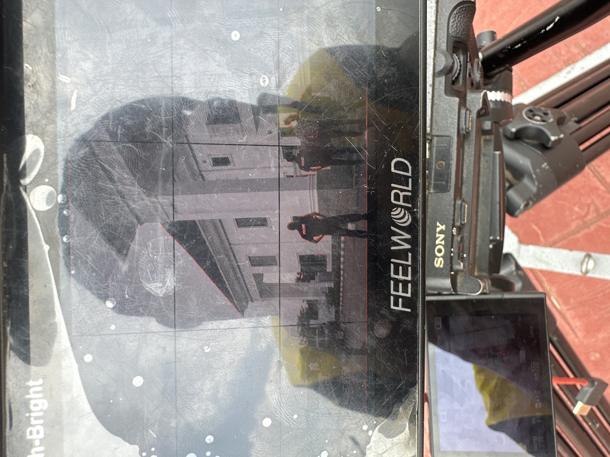 The image shows a close-up of a black monitor screen with a reflection of a building and several people. The monitor displays the brand "FEELWORLD" and "SONY" is visible on the camera below. The building appears to be a light-colored, possibly white, structure with windows and a distinct architectural style. In the reflection, several individuals are visible standing in front of the building. Some appear to be wearing dark uniforms with "POLICE" visible on the back of one. The overall scene suggests a filming or photography setup, possibly documenting an event or location. The environment outside the immediate focus appears to be outdoors with a cloudy sky reflected in the monitor. The location in Pakuro, Nigeria is not explicitly identifiable from the image itself, but the presence of police uniforms might suggest a public or official area. The monitor screen has numerous scuff marks and some circular smudges, indicating it might be used in various conditions. The camera below is mounted on a tripod, further emphasizing the professional recording context.