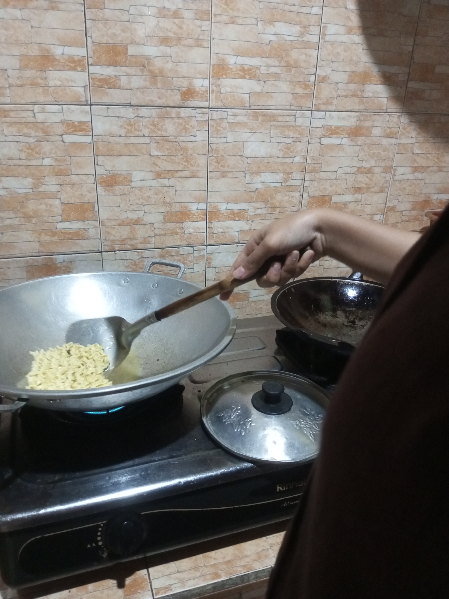 The image shows a person cooking instant noodles in a wok on a gas stove. The background is a tiled wall. The scene appears to be in a kitchen. The gas stove is black with a knob that has markings and dots. A metal lid with a black handle is placed on the stove next to the wok. The wok contains a portion of uncooked instant noodles and oil. The person's hand is visible, holding a spatula and stirring the noodles. Another pan is visible to the right of the wok. The overall impression is of a domestic cooking scene.
