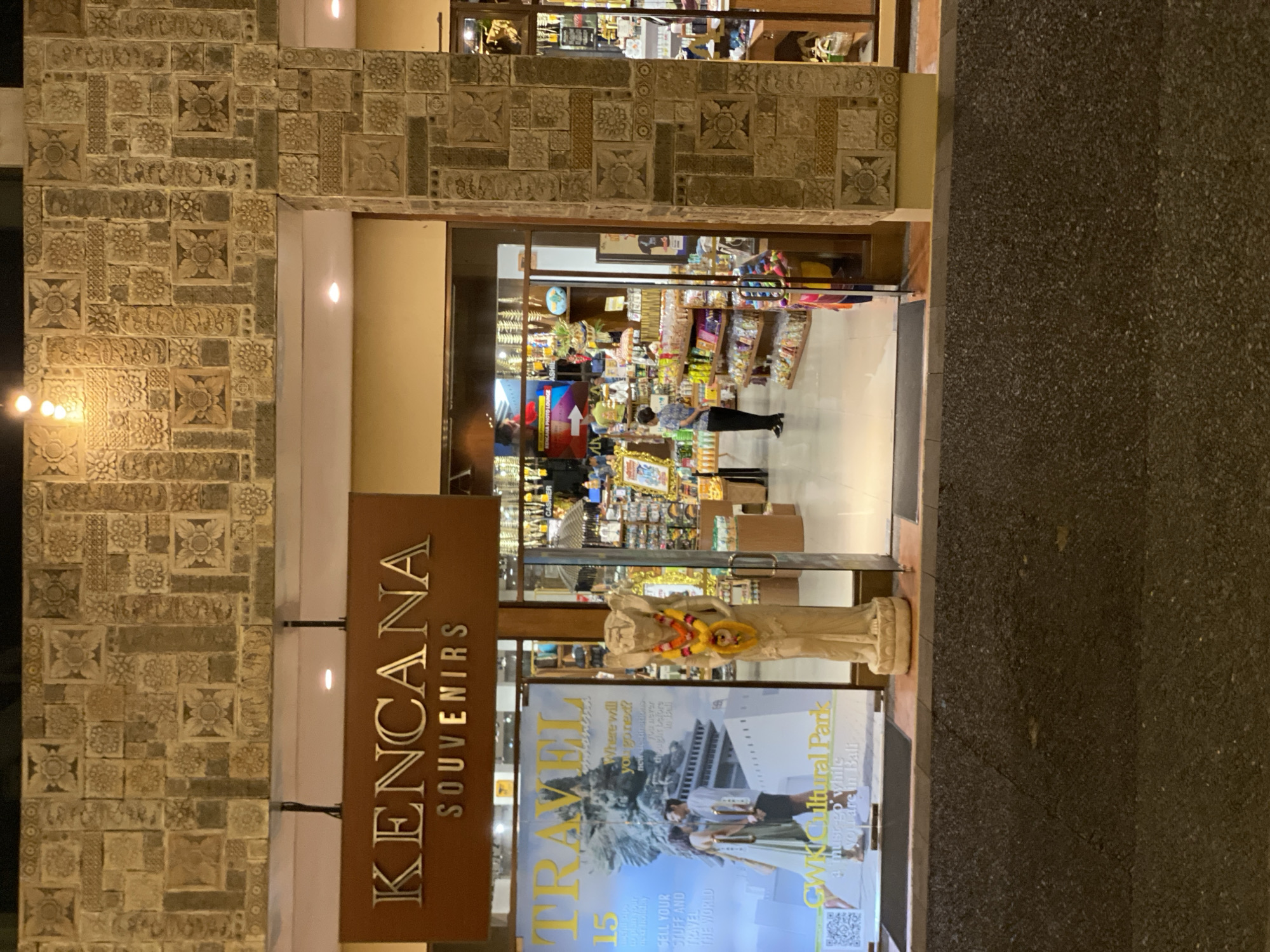 The image depicts the entrance to a souvenir shop named "KENCANA SOUVENIRS" in Kuta Selatan, Indonesia. The shop's facade features traditional Balinese stone carvings on its upper walls and columns. In front of the entrance, a statue adorned with a lei stands next to a large magazine display. The magazine features a cover story titled "TRAVEL" and highlights GWK Cultural Park. Inside the store, shelves are stocked with various souvenirs and products, and a cashier is visible at the back. A woman in a blue shirt and black pants is standing near the entrance, facing into the store. The ground in front of the shop is paved with dark asphalt, and there is a subtle curb separating it from the shop's entrance. The lighting suggests it is evening or nighttime, with interior lights illuminating the shop.