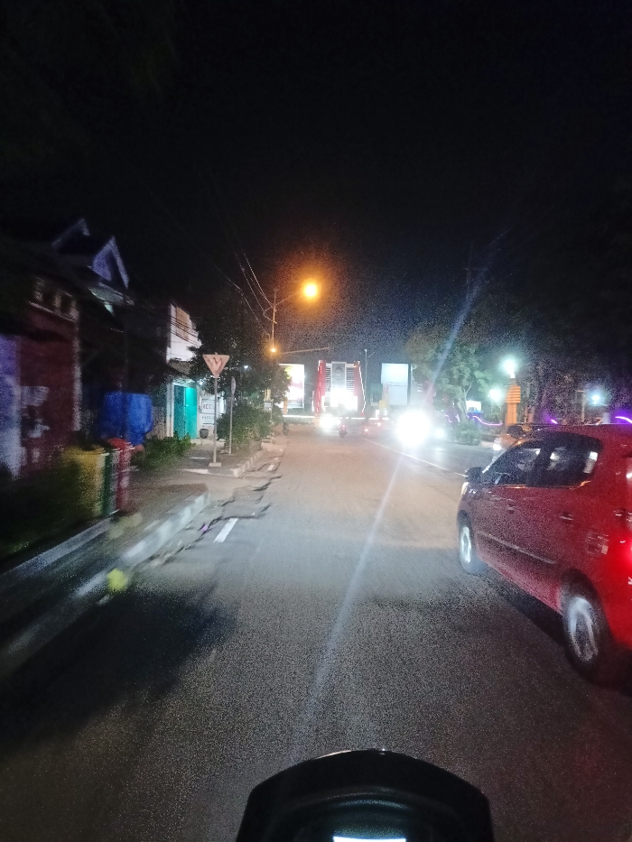 A night-time street scene is depicted from the perspective of a vehicle, likely a motorcycle or scooter, as suggested by the dashboard visible at the bottom. The sky is dark, indicating night, with various artificial light sources illuminating the area. This scene is set in Mojosari, Indonesia.
On the left side of the road, several buildings, possibly residential or commercial, line the street, some displaying internal or external lights. A sidewalk with visible wear and tear runs alongside the buildings. Further down the street on the left, a triangular road sign, likely a 'yield' or 'give way' sign, stands next to a pole with power lines. A bright yellow-orange streetlight casts a glow near the middle-left of the frame.
The asphalt road surface appears slightly reflective, possibly damp. A white line is painted on the road, near the sidewalk. On the right side of the frame, a red four-door car is visible, its tail lights illuminated, either moving or parked. In the distance, on the road ahead, extremely bright headlights from an oncoming vehicle (possibly a truck or bus given its height) create a strong glare that obscures details. Another vehicle with bright headlights is further behind it.
The activity taking place is travel along a street at night. No people are clearly visible. No text is clearly legible on any signs or objects within the image. The overall impression is of urban or suburban travel at night.