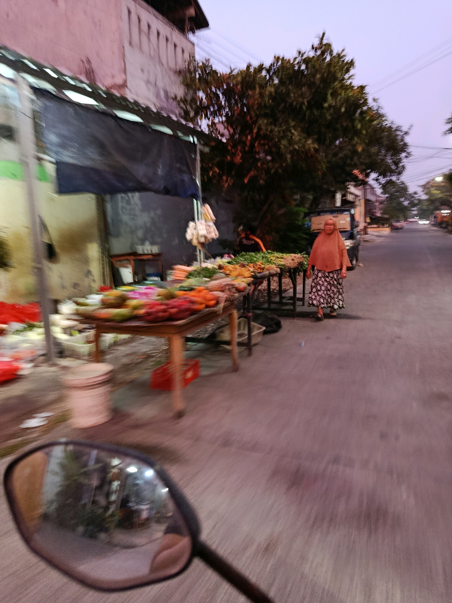 A blurred, eye-level shot captures a street scene at dusk in Bekasi, Indonesia. A woman wearing a hijab and a patterned skirt walks down the right side of the asphalt road, facing away from the camera. She is seen between two tables laden with fresh produce on the left, suggesting a street market. One table, closer to the camera, displays an assortment of fruits, including what appear to be dragon fruit, mangoes, and possibly rambutans, in various colors like red, yellow, and green. Behind this table, another table is visible with more vegetables and fruits. A building with a pinkish facade and white window frames is situated to the far left, partially obscured by a dark tarp. Trees with dense foliage are present behind the market stalls, and power lines crisscross the pale purple sky. In the foreground, the reflection of the scene is visible in the rounded mirror of a motorcycle, indicating the viewpoint of the photographer. The motion blur suggests movement, likely from the motorcycle. A white bucket and a red crate are on the ground near the market stalls. In the background, other vehicles and the continuation of the street market are faintly discernible.